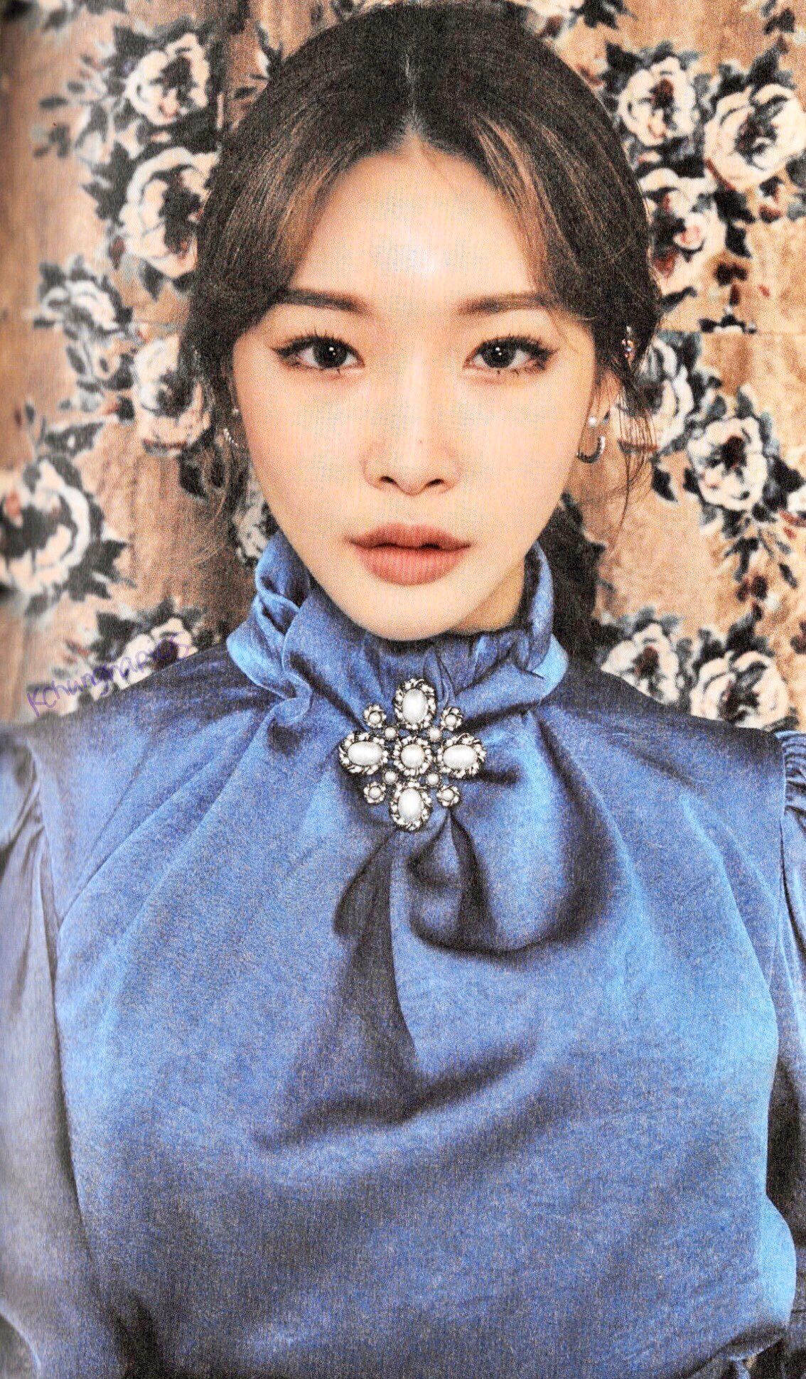 Chungha 桌布 壁紙 — 一位身穿藍色高領襯衫、佩戴鑲寶石胸針的女性，在花卉壁紙背景下向前凝視。