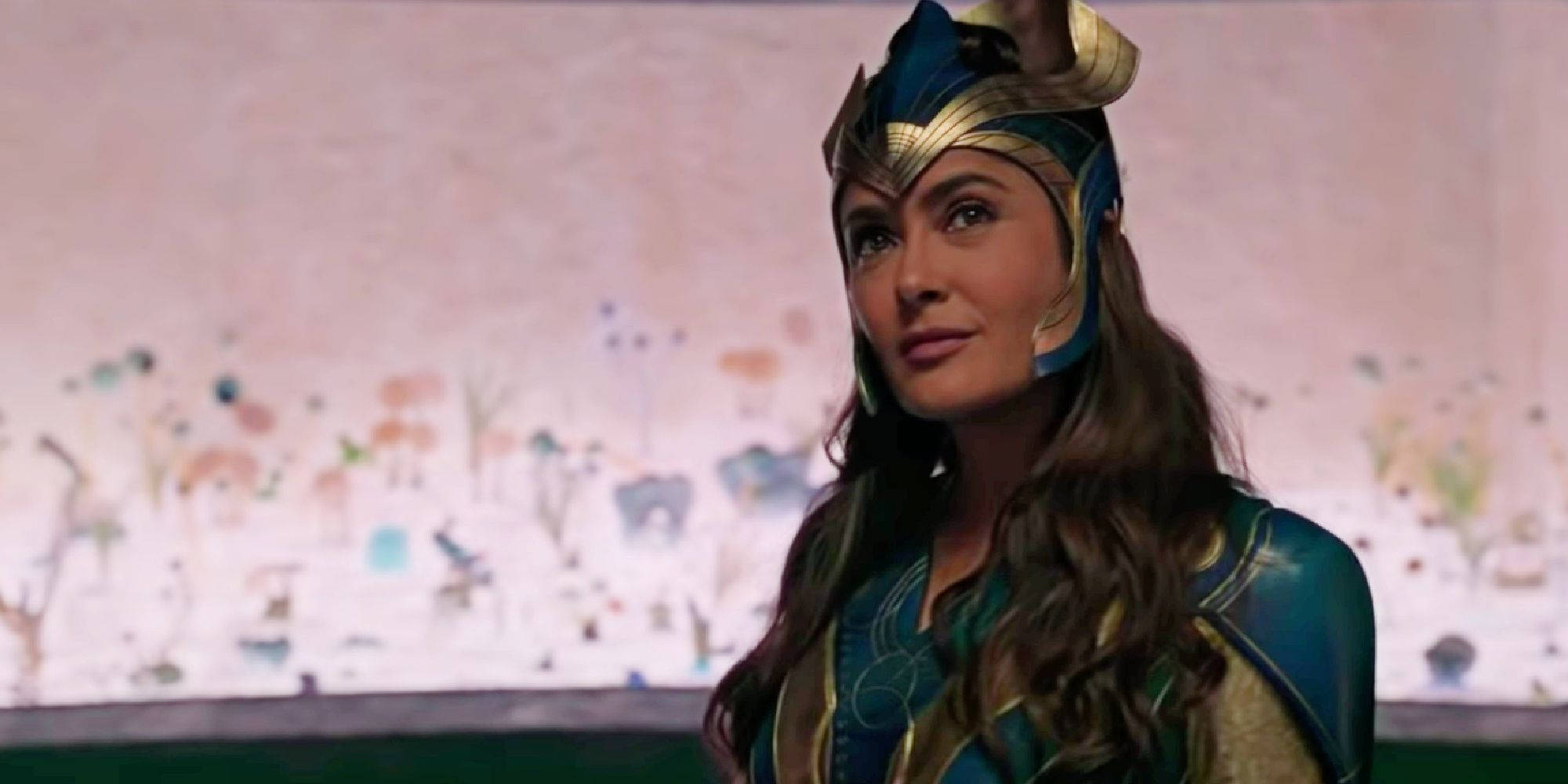 Eternals 桌布 壁紙 — 一位身穿藍金色盔甲、戴著頭盔的女性站在彩繪牆前。