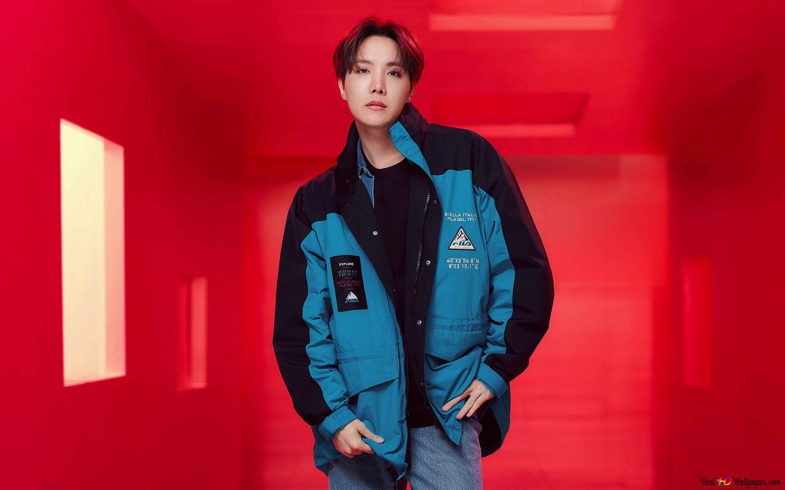 Fila 桌布 壁紙 — 一位身穿藍黑外套的年輕男子自信地站在一條帶有發光矩形窗戶的鮮紅色走廊中。