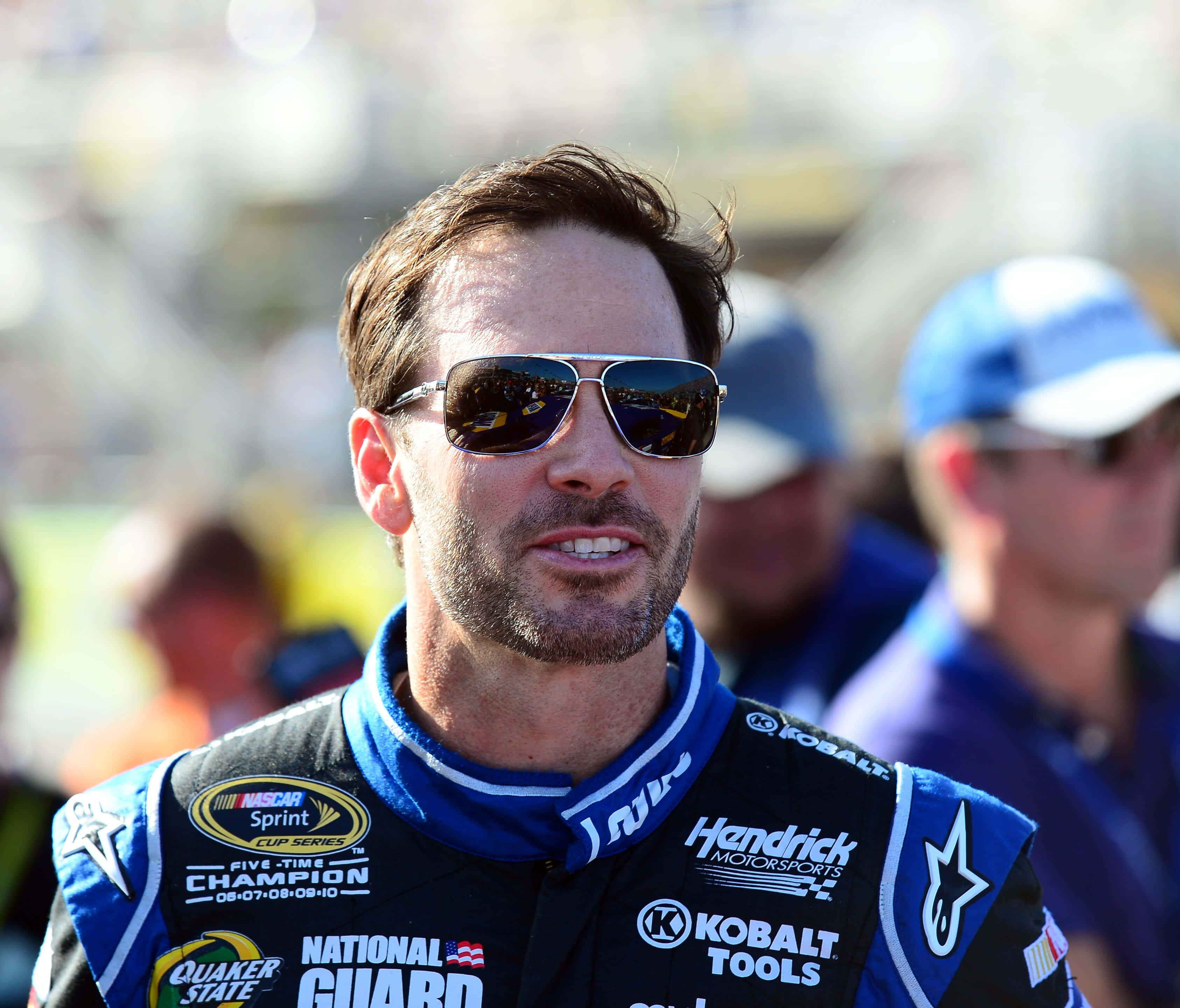 Jimmie Johnson 桌布 壁紙 — 一位身穿藍黑賽車服、帶有贊助商標誌的NASCAR車手，在明亮的日光下自信地站著，戴著飛行員太陽眼鏡。