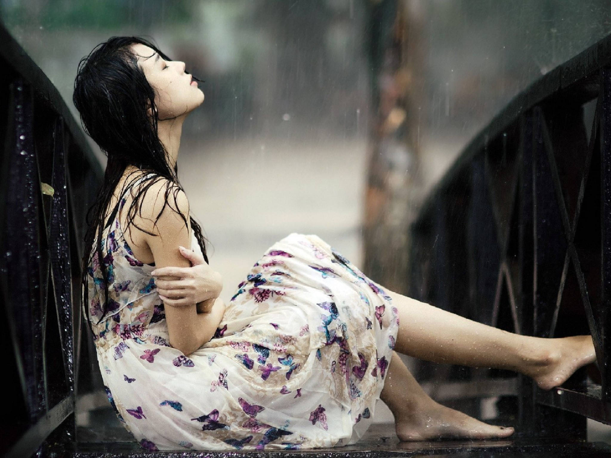 Sad Girl 桌布 壁紙 — 一位身穿蝴蝶印花連衣裙的女子赤腳坐在濕漉漉的橋欄杆上，頭向後仰，雨水在她周圍落下。