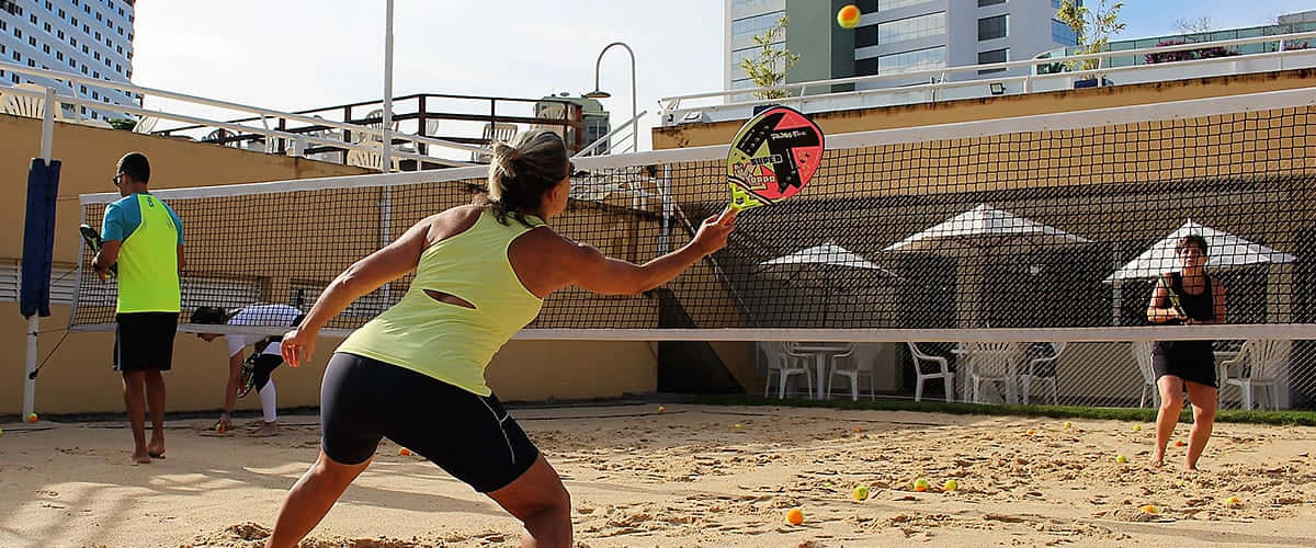 Beach Tennis 桌布 壁紙 — 一位身穿螢光黃上衣的女性在陽光普照的屋頂沙地球場上揮拍擊打網球，背景是城市建築。