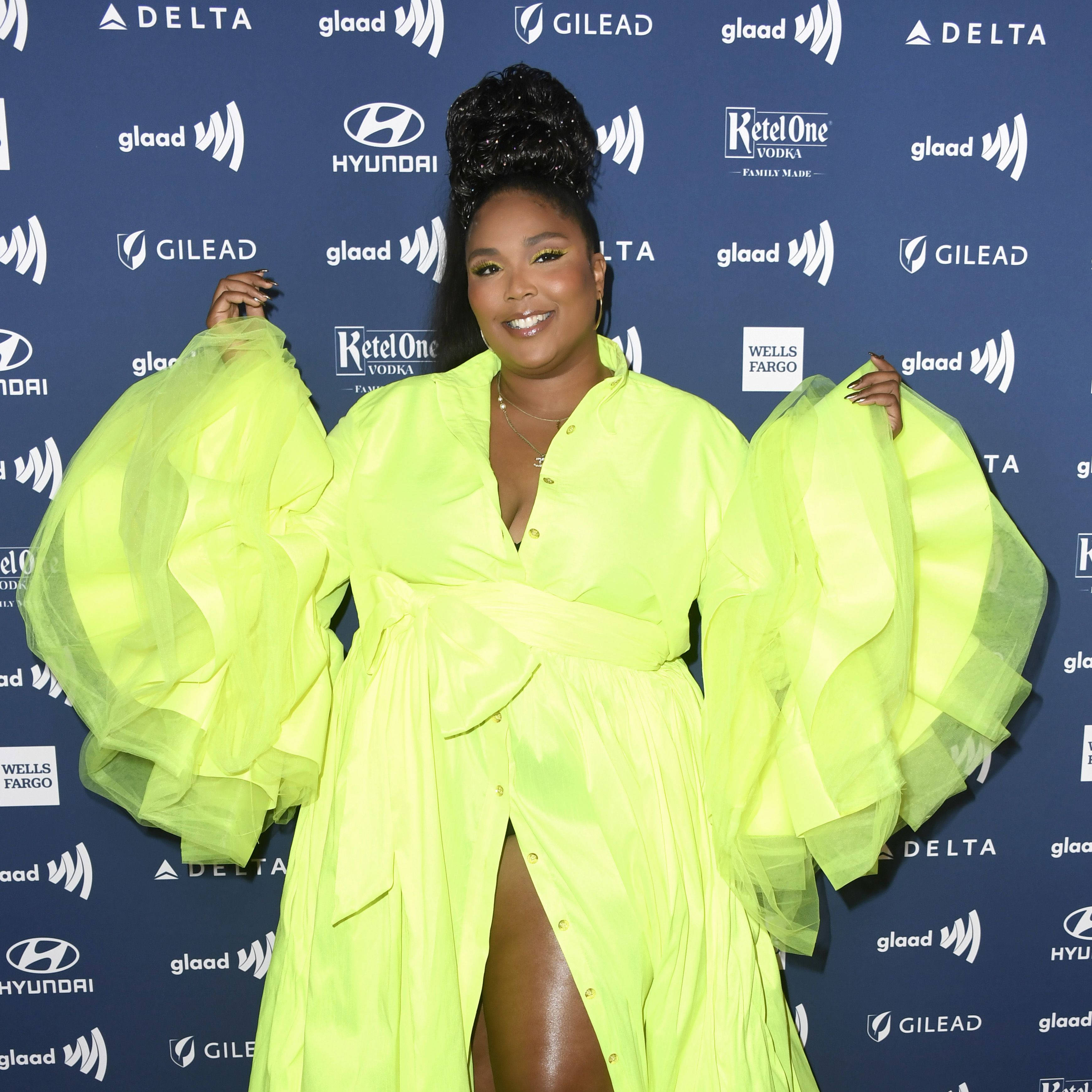 Lizzo 桌布 壁紙 — 一位身穿螢光黃色禮服、袖子蓬鬆的耀眼女性，在活動中自信地站在品牌藍色背景前擺姿勢。