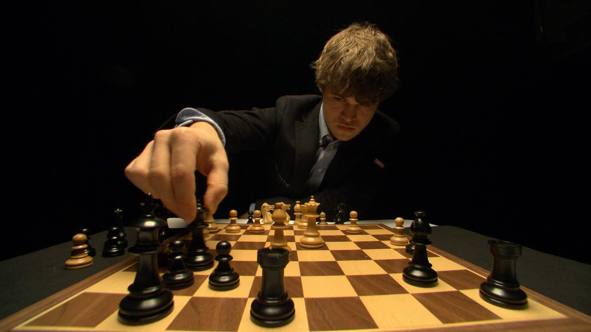 Magnus 桌布 壁紙 — 一位身穿西裝、專注的西洋棋棋手在黑色背景的戲劇性聚光燈下，在木製棋盤上移動黑兵。