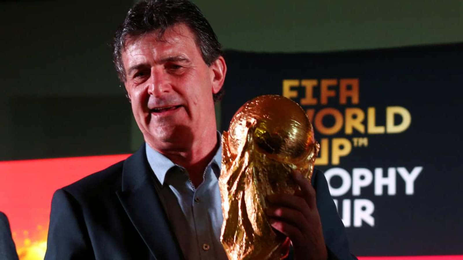Trophy 桌布 壁紙 — 一位身穿西裝、面帶微笑的男子，在品牌背景前自豪地舉起閃閃發光的金色FIFA世界盃獎盃。