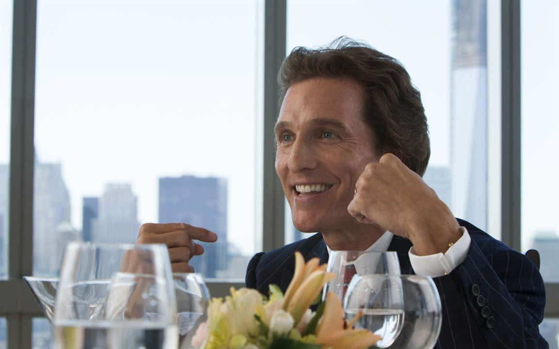 Wolf Of Wall Street 桌布 壁紙 — 一位身穿西裝、面帶微笑的男子，在可俯瞰城市天際線的餐桌上舉拳慶祝。