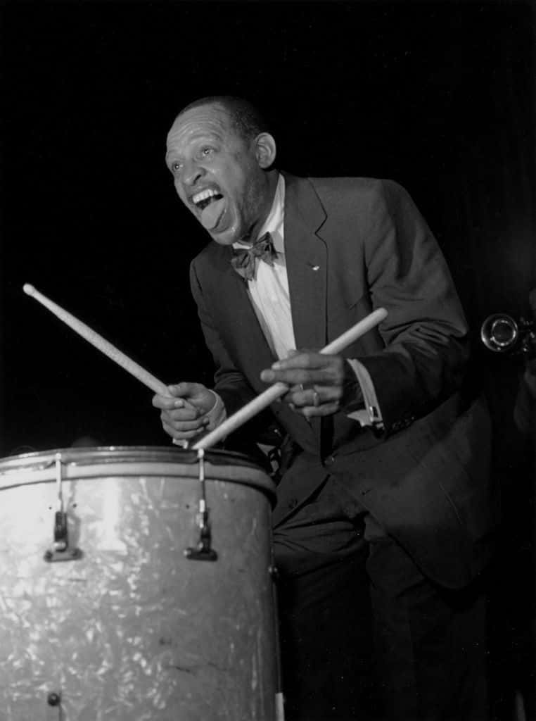 drums 桌布 壁紙 — 一位身穿西裝和蝴蝶結的快樂爵士鼓手，在黑白照片中手持鼓棒放在低音鼓上，狂野大笑。