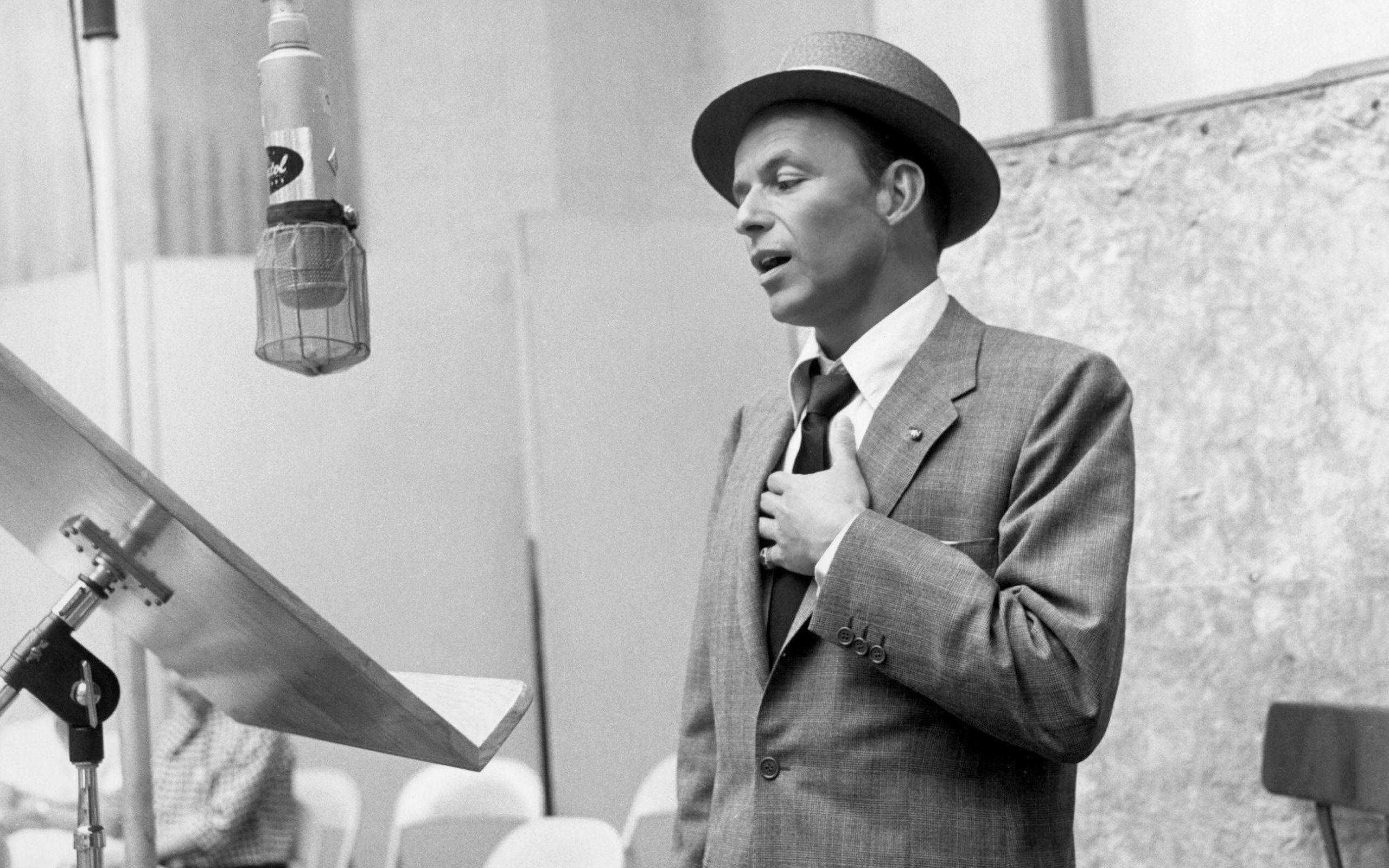 Sinatra 桌布 壁紙 — 一位身穿西裝和費多拉帽的男子在錄音室中熱情地對著復古麥克風唱歌。