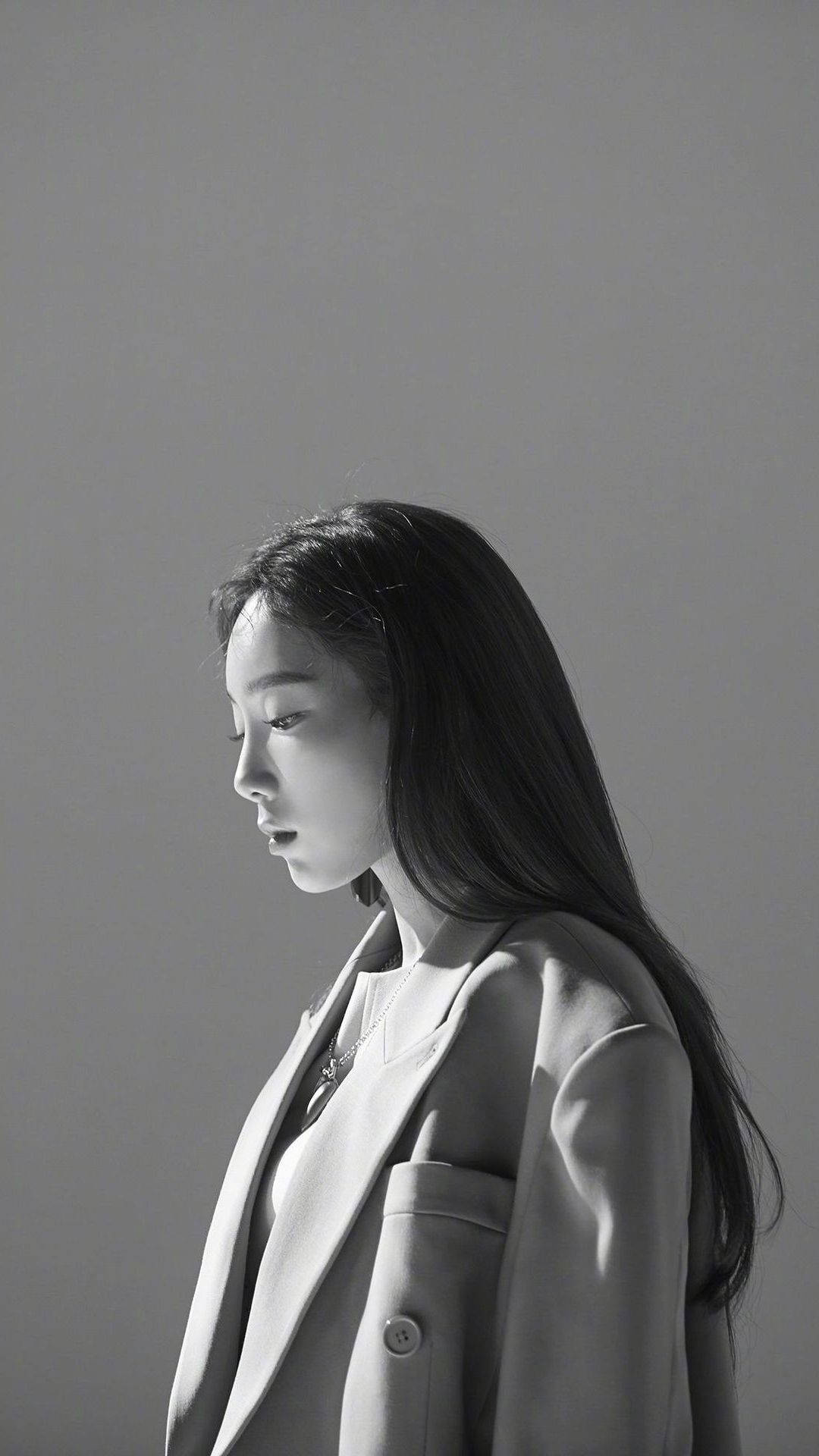 Taeyeon 桌布 壁紙 — 一位身穿西裝外套的女性站在柔和光線中，背景為純灰色，她目光低垂，陷入靜默沉思。