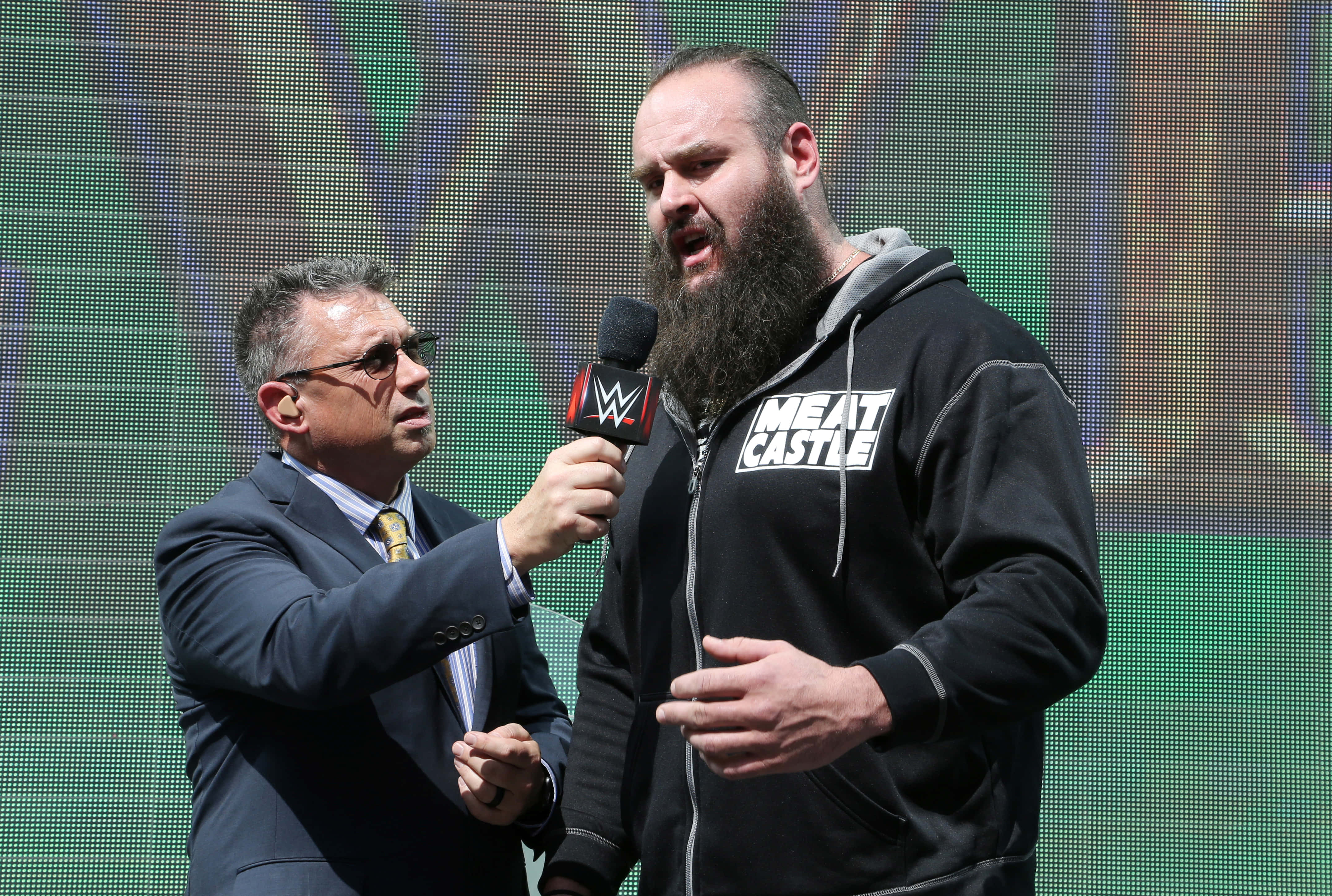 WWE 桌布 壁紙 — 一位身穿西裝的WWE記者，在現場活動中將麥克風對準一位穿著黑色「MEAT CASTLE」連帽衫的鬍鬚摔角手。