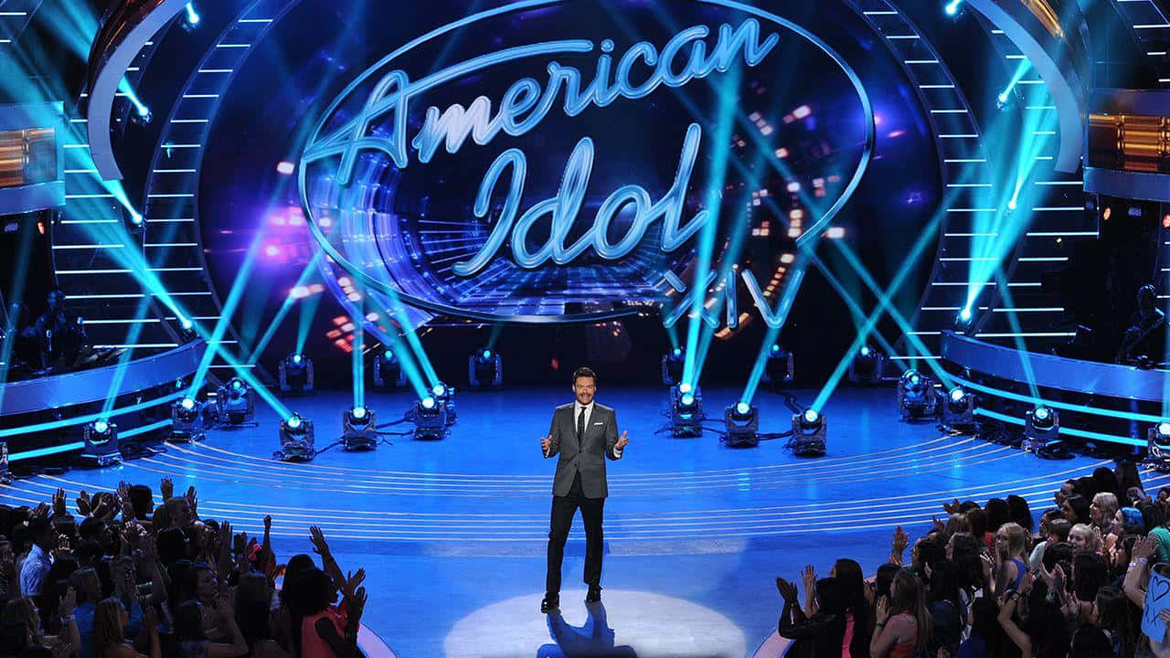 american idol 桌布 壁紙 — 一位身穿西裝的主持人站在舞台中央，在明亮的藍色燈光下，面對著美國偶像現場演出中歡呼的觀眾。