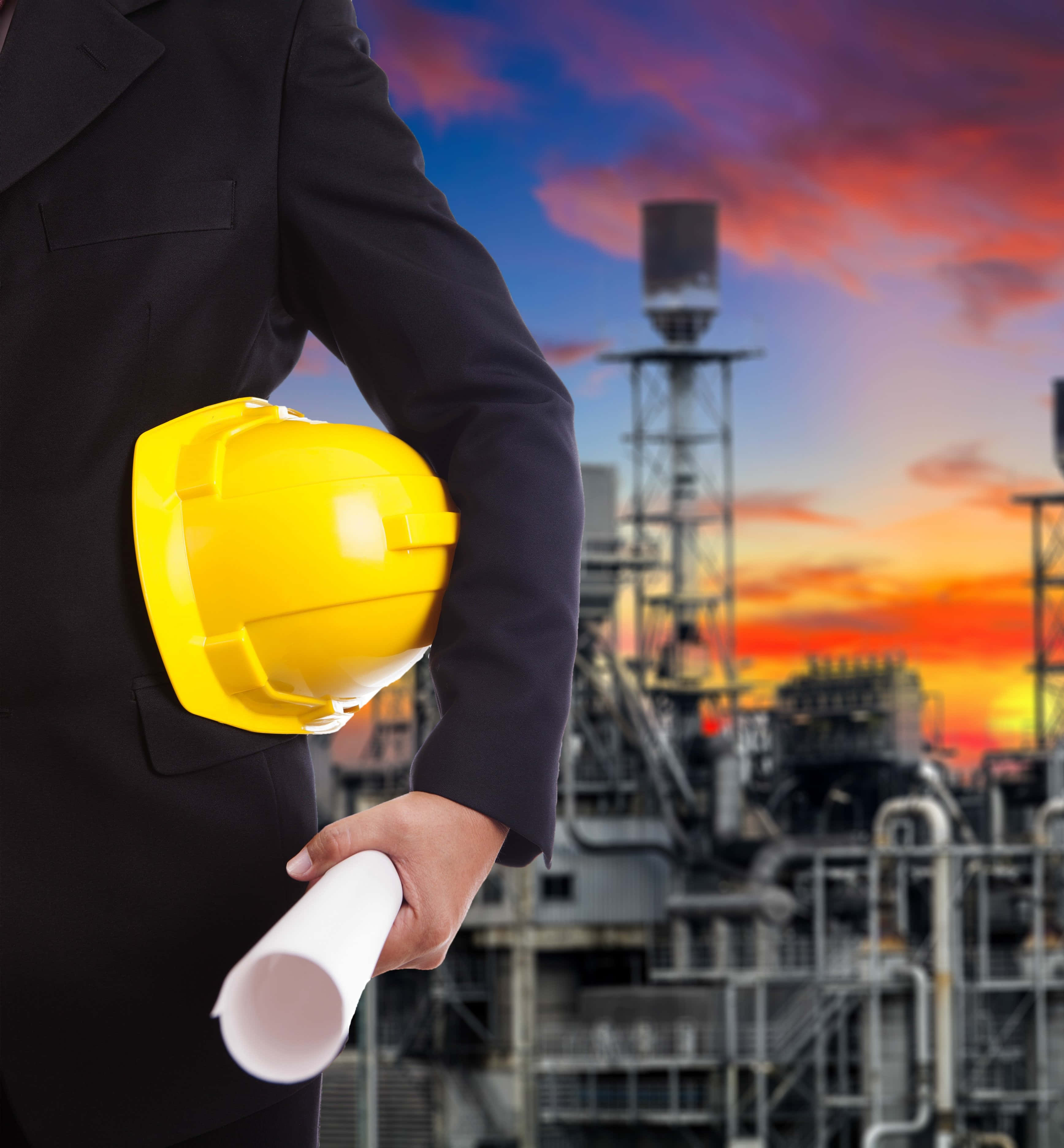 construction 桌布 壁紙 — 一位身穿西裝的商人手持黃色安全帽和藍圖，背景是夕陽下的工業廠房。