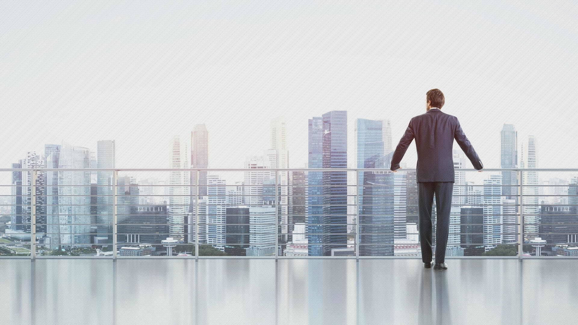 商業 桌布 壁紙 — 一位身穿西裝的商人站在玻璃陽台上，俯瞰著明亮天空下高樓林立的現代城市天際線。
