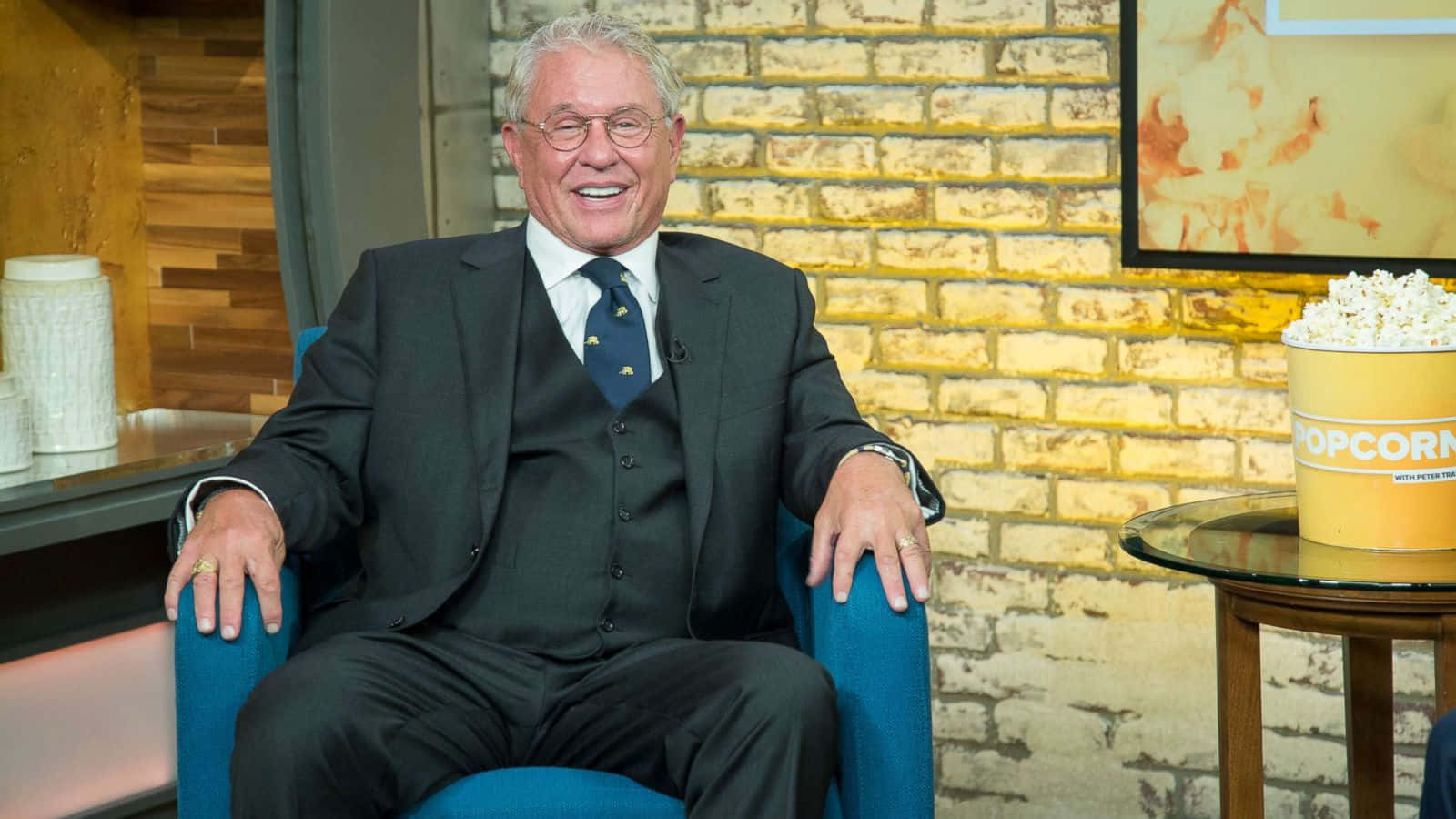 Tom Berenger 桌布 壁紙 — 一位身穿西裝的年長男士坐在藍色扶手椅上，旁邊放著一桶爆米花，開懷大笑。