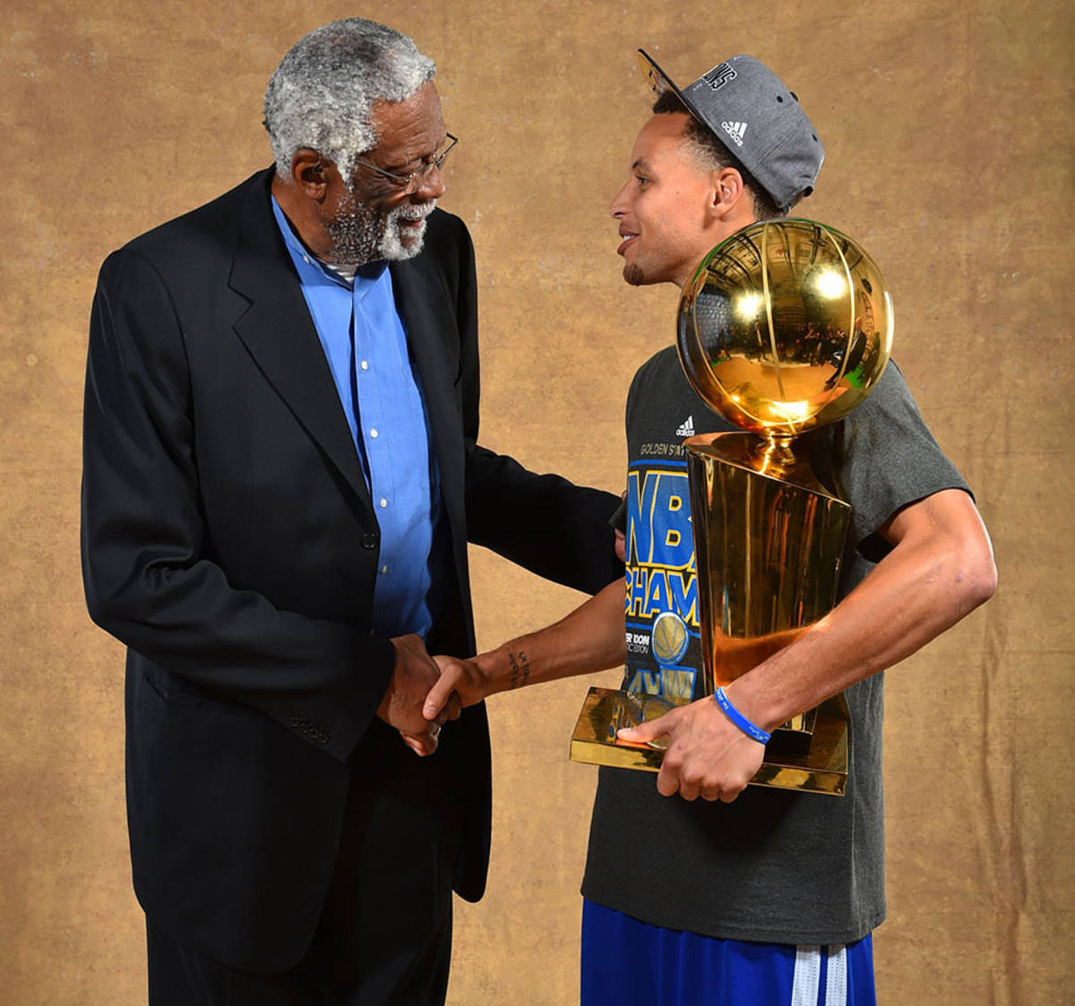 NBA 桌布 壁紙 — 一位身穿西裝的年長男子與一位年輕籃球運動員握手，後者手持閃亮的NBA冠軍獎盃，背景為溫暖的棕色。