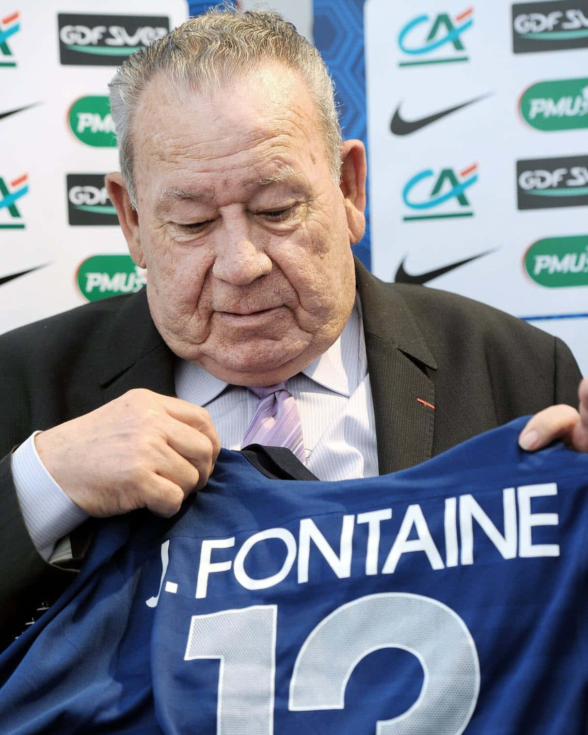 足球 桌布 壁紙 — 一位身穿西裝的長者在記者會上舉起一件印有「J FONTAINE 12」的藍色足球球衣。