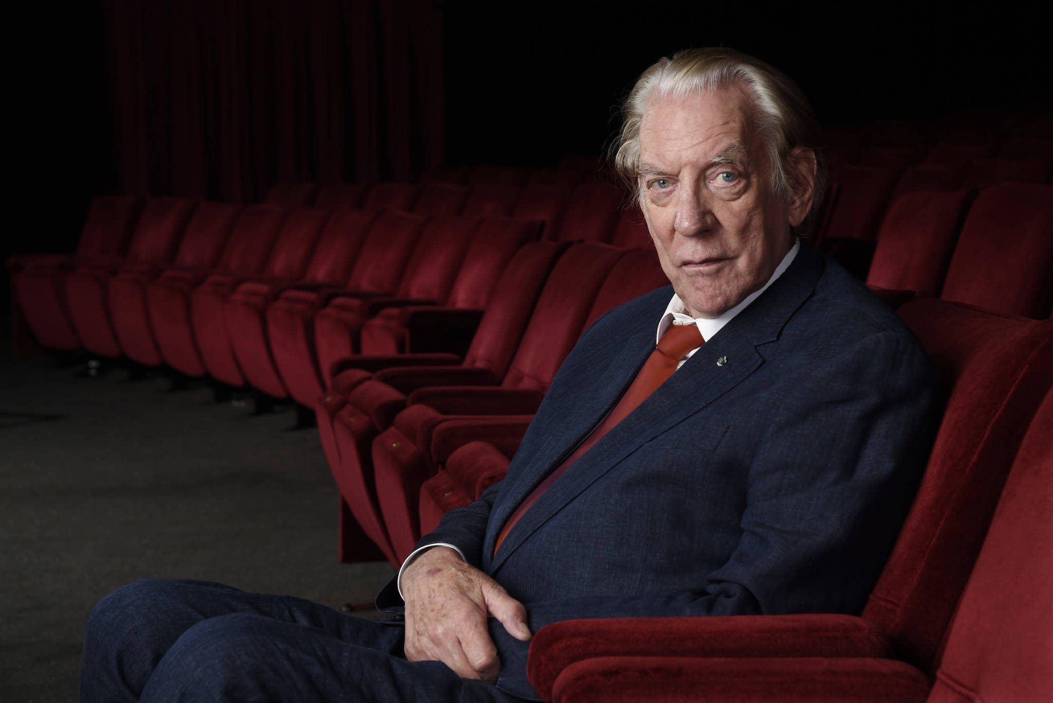 actor 桌布 壁紙 — 一位身穿西裝的長者獨自坐在燈光昏暗的劇院裡，周圍是一排排紅色天鵝絨座椅。