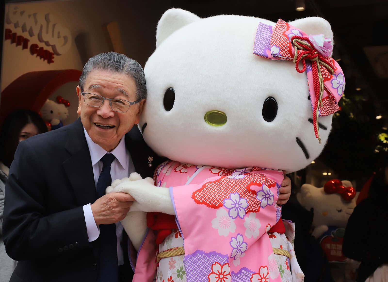 Hello Kitty 桌布 壁紙 — 一位身穿西裝的長者笑容滿面，擁抱著一個穿著粉紅色花卉和服、繫著圖案蝴蝶結的巨型Hello Kitty吉祥物。