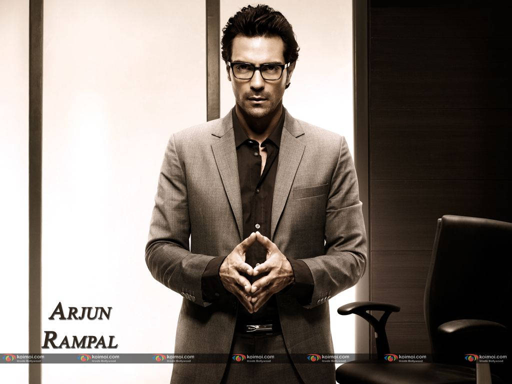 Arjun Rampal 桌布 壁紙 — 一位身穿訂製西裝和眼鏡的男子，雙手交握，自信地站在現代辦公室中，沐浴在柔和的工作室燈光下。