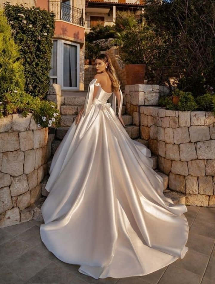 Wedding Gown 桌布 壁紙 — 一位身穿豐盈緞面婚紗的新娘，優雅地站在石階上，四周環繞著茂密的綠植和溫暖的陽光。