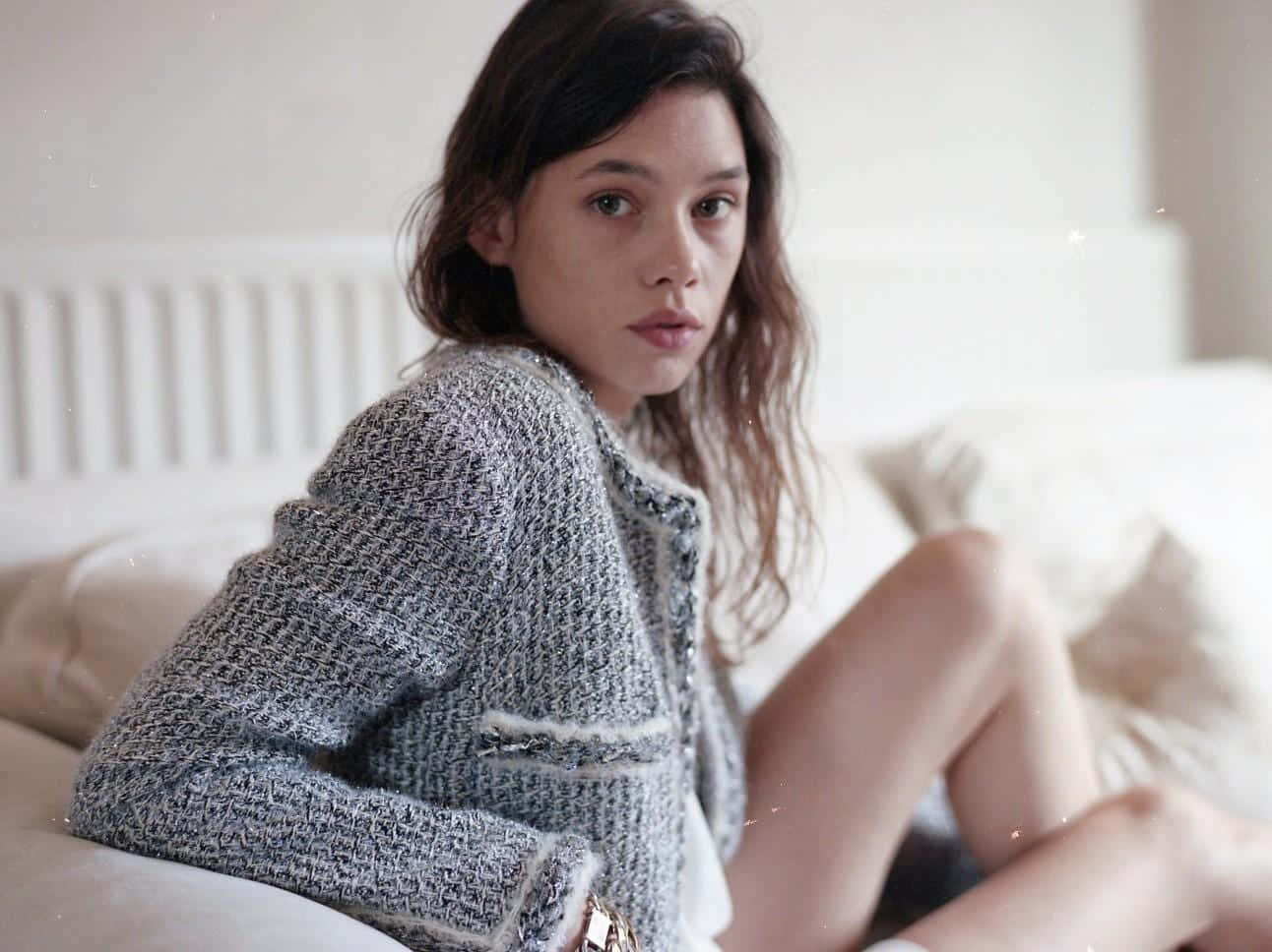 Astrid Berges Frisbey 桌布 壁紙 — 一位身穿質感灰色外套的年輕女性坐在白色床上，直視鏡頭，表情柔和而中性。