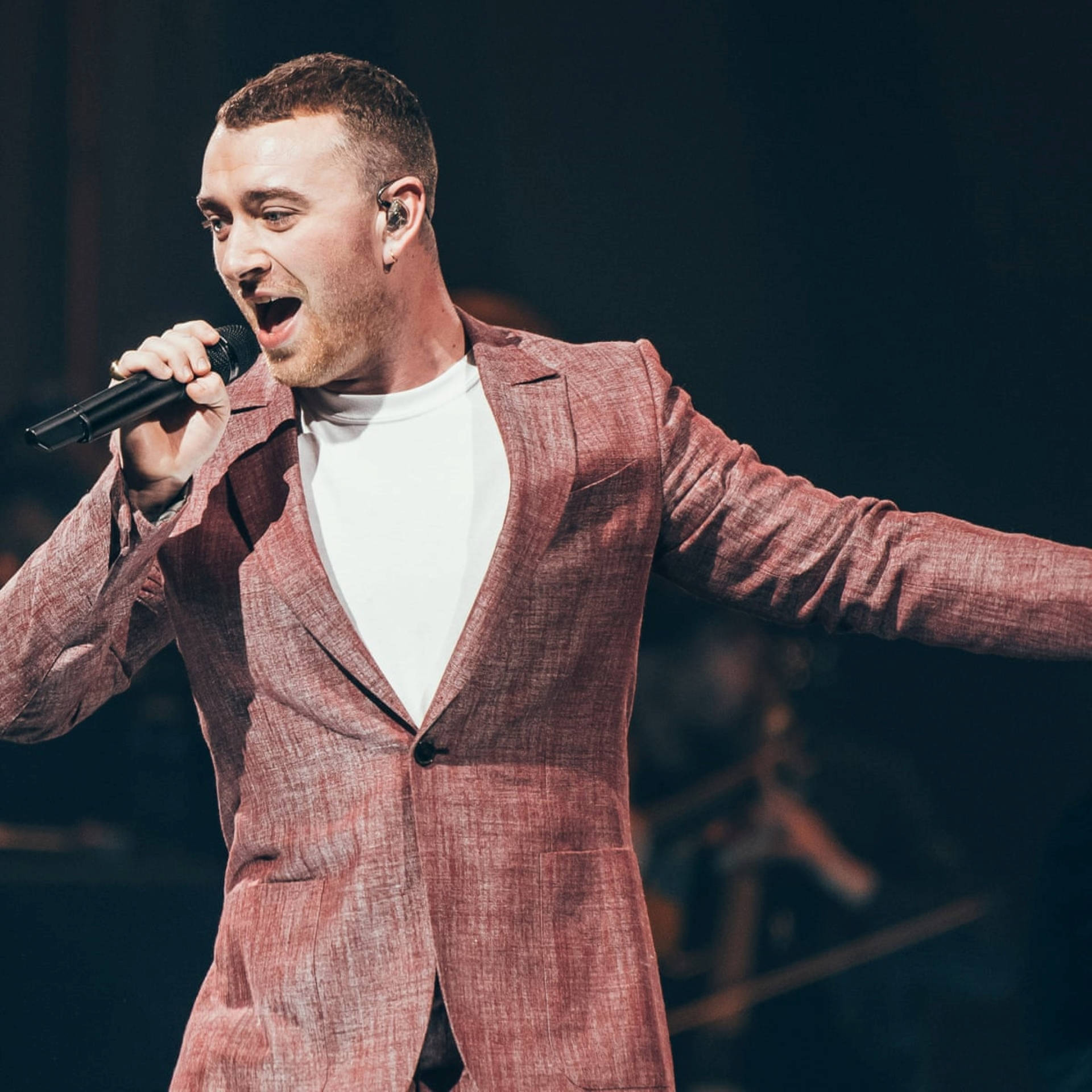 Sam Smith 桌布 壁紙 — 一位身穿質感粉紅色西裝的男歌手在戲劇性的舞台燈光下張開雙臂熱情表演。
