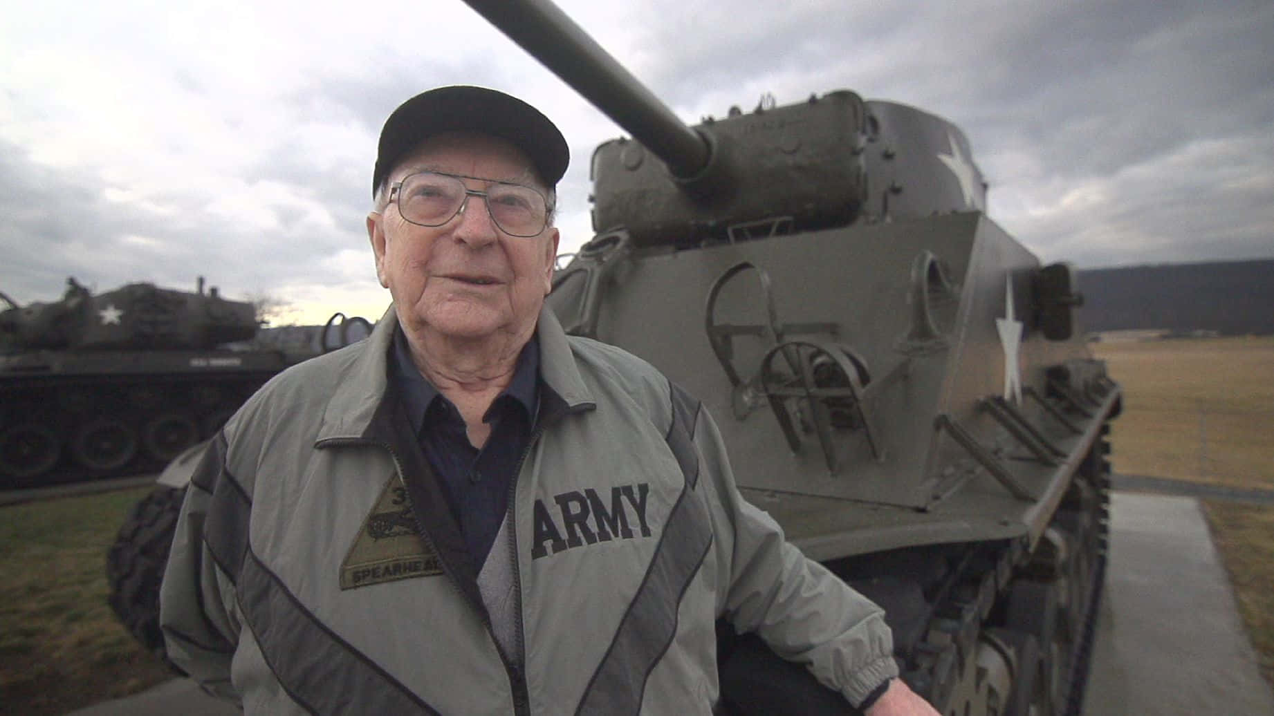 Ww2 桌布 壁紙 — 一位身穿軍裝夾克的老年退伍軍人，在陰天自豪地站在軍用坦克前。