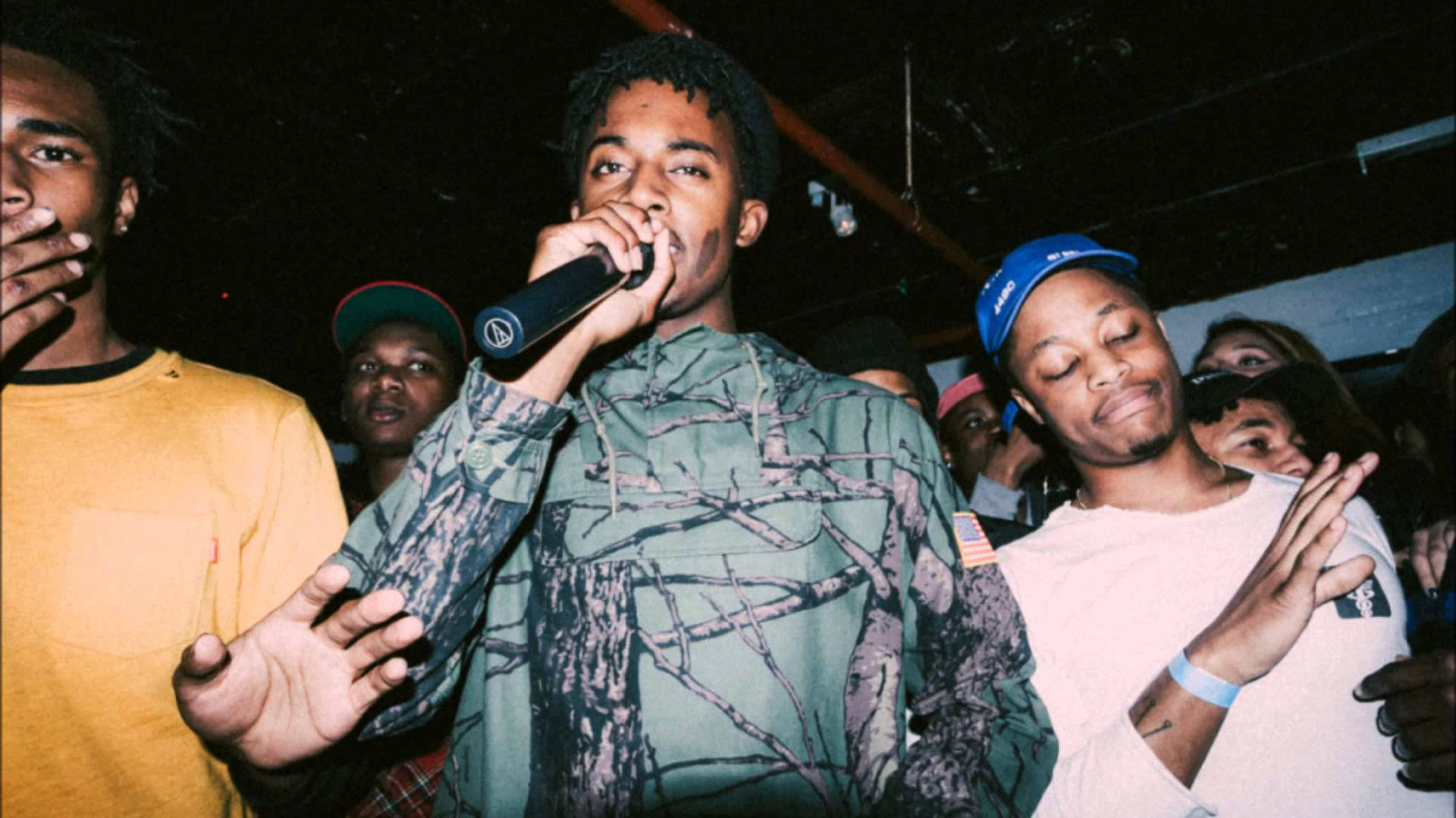 Carti 桌布 壁紙 — 一位身穿迷彩連帽衫的年輕男子在昏暗的場地中，對著麥克風說唱，周圍是投入的群眾。