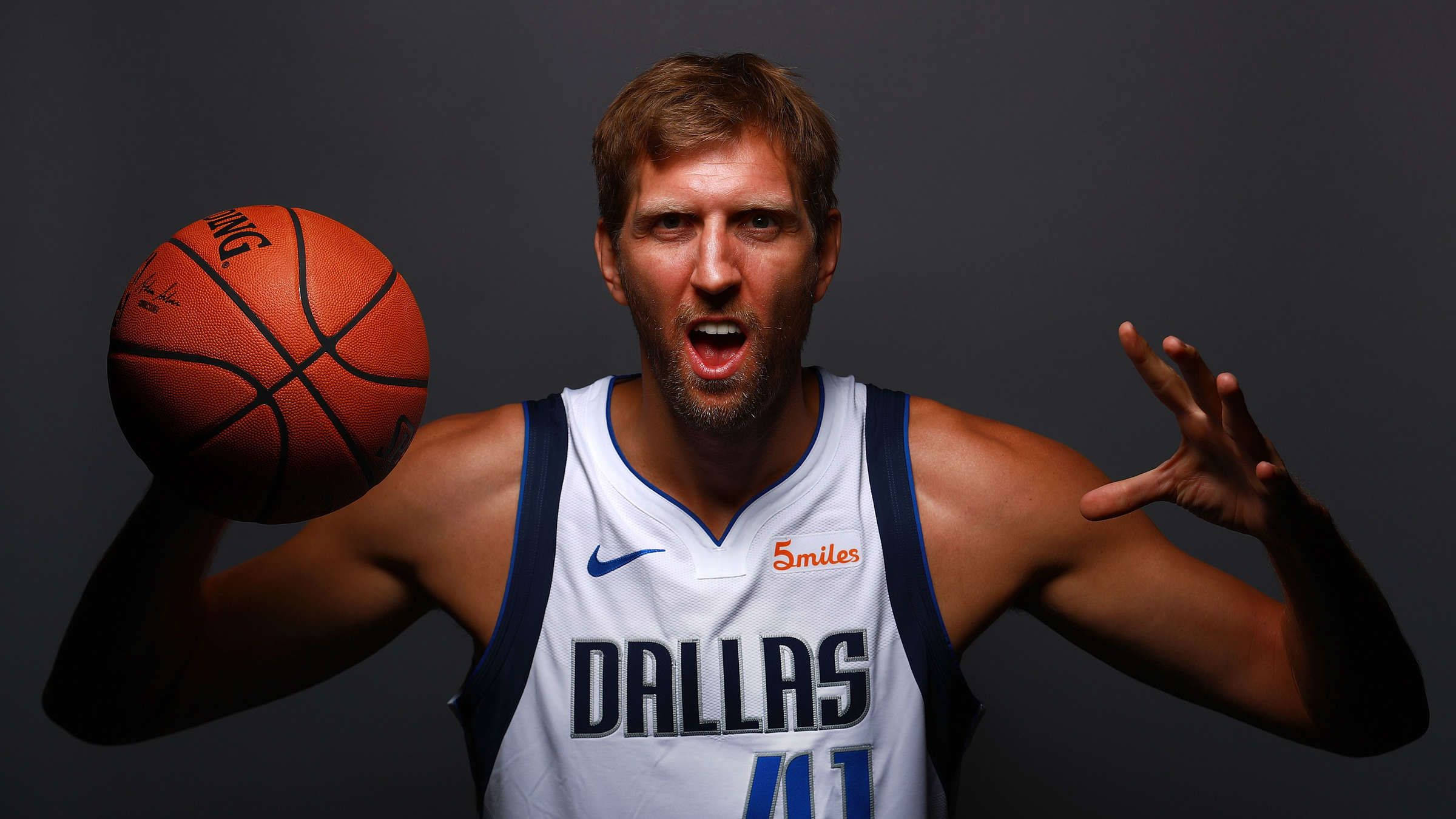 Dirk Nowitzki 桌布 壁紙 — 一位身穿達拉斯球衣的籃球運動員在黑暗的攝影棚背景下拿著球並做出激烈的姿勢。