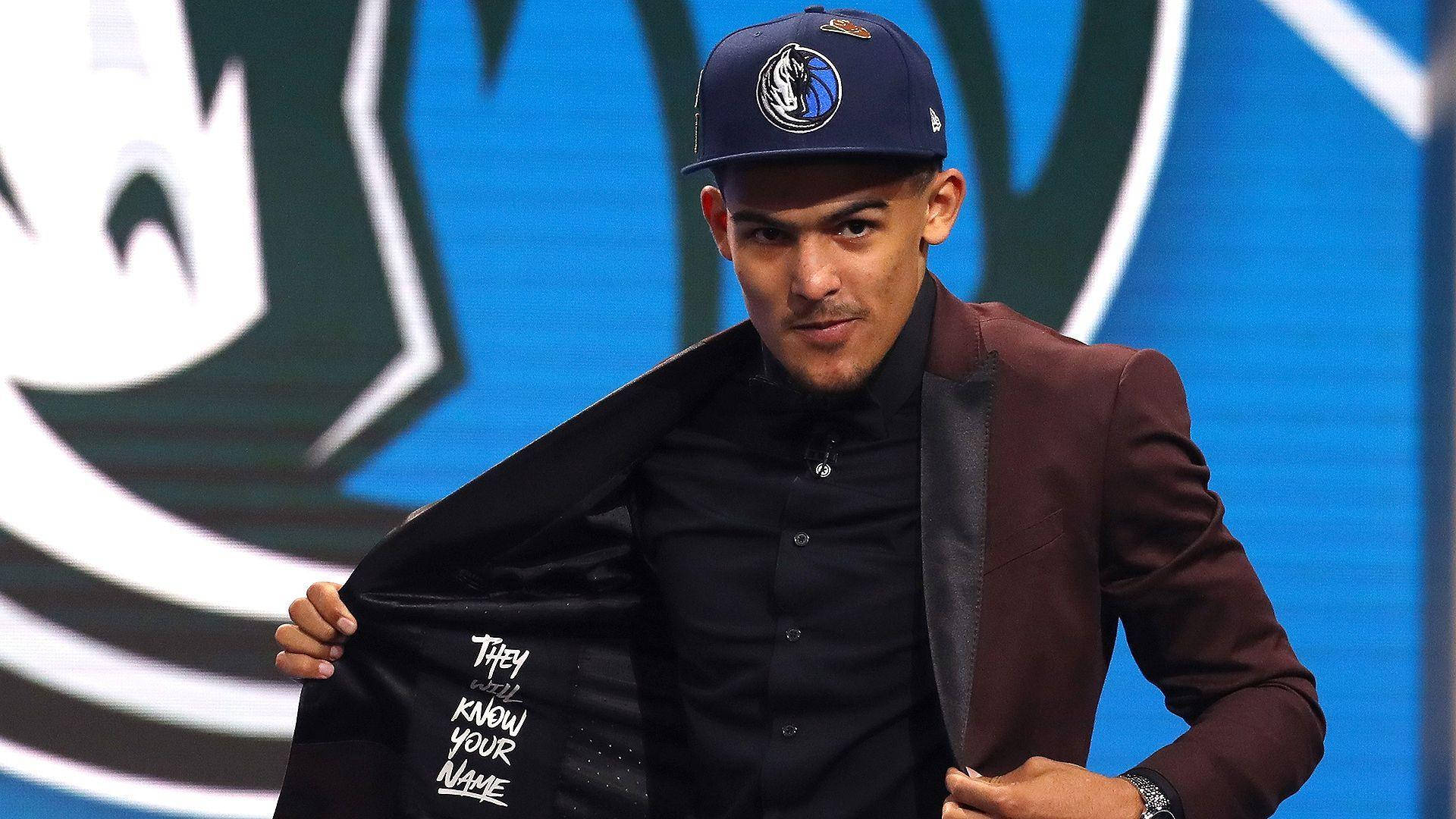 Trae Young 桌布 壁紙 — 一位身穿酒紅色西裝外套和達拉斯獨行俠帽子的籃球運動員，戲劇性地揭開一件寫有「They Know Your Name」的外套。