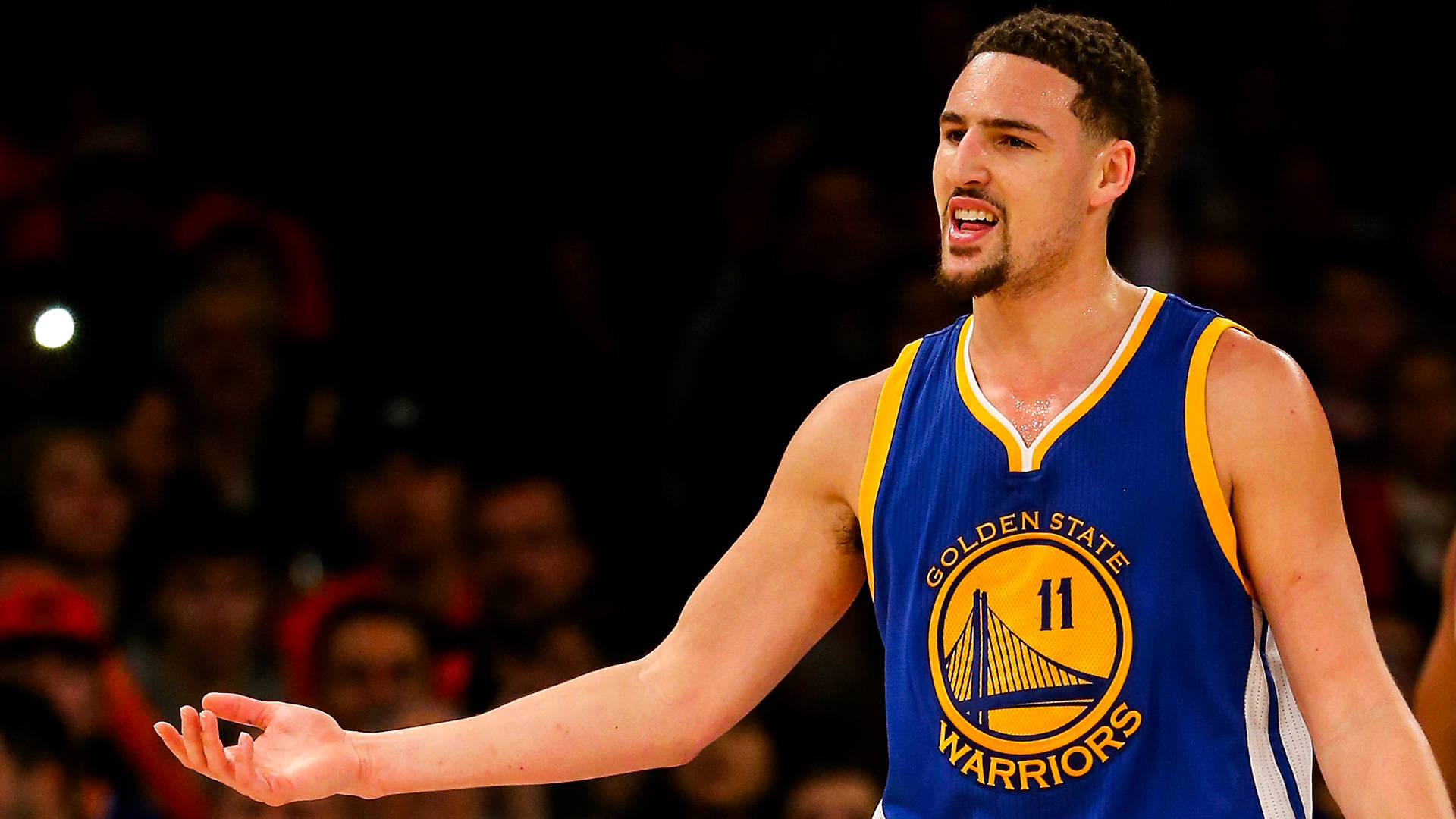 Thompson 桌布 壁紙 — 一位身穿金州勇士隊球衣的籃球運動員在明亮的場館燈光下比賽時激動地做手勢。