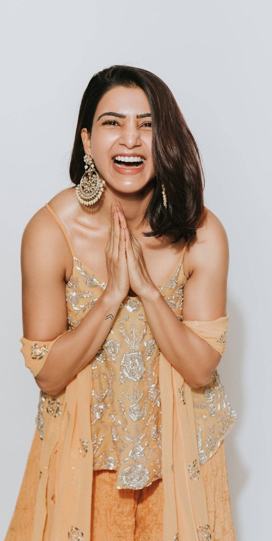 Celebrity 桌布 壁紙 — 一位身穿金色刺繡服裝的快樂女性，在純白背景前雙手合十大笑。