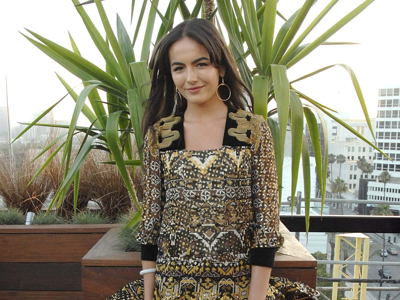 Camilla Belle 桌布 壁紙 — 一位身穿金色和黑色圖案連衣裙的女性微笑著靠在一個種有高大綠色植物的屋頂花盆上，身後是城市天際線。