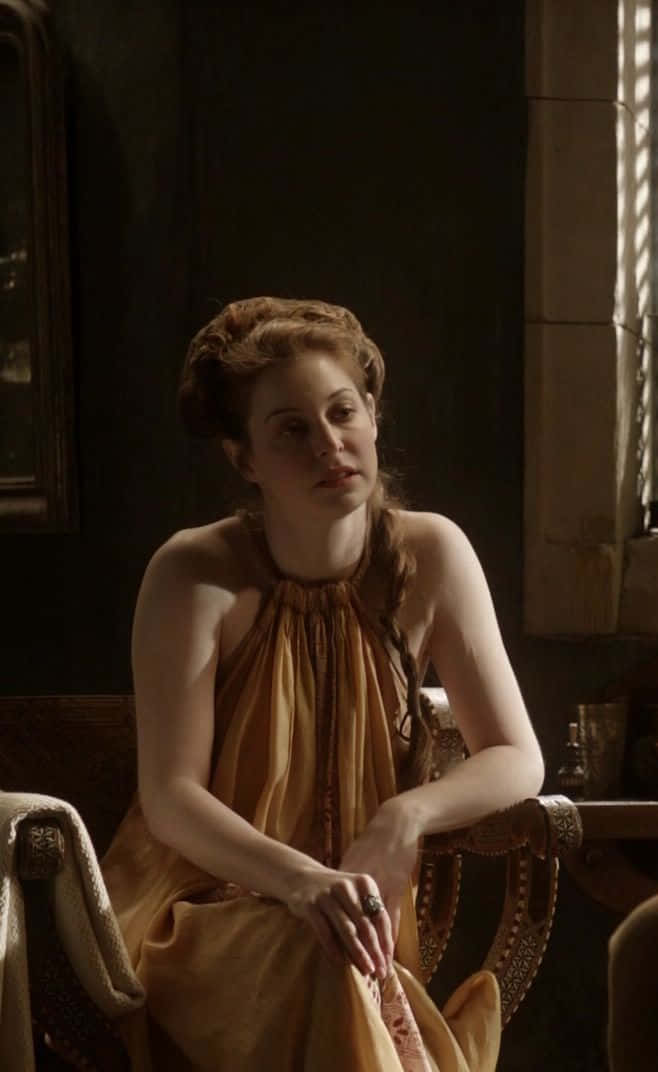 Esmé Bianco 桌布 壁紙 — 一位身穿金色垂墜長袍的女子坐在陽光普照的華麗椅子上，在深色石牆前若有所思地凝視。