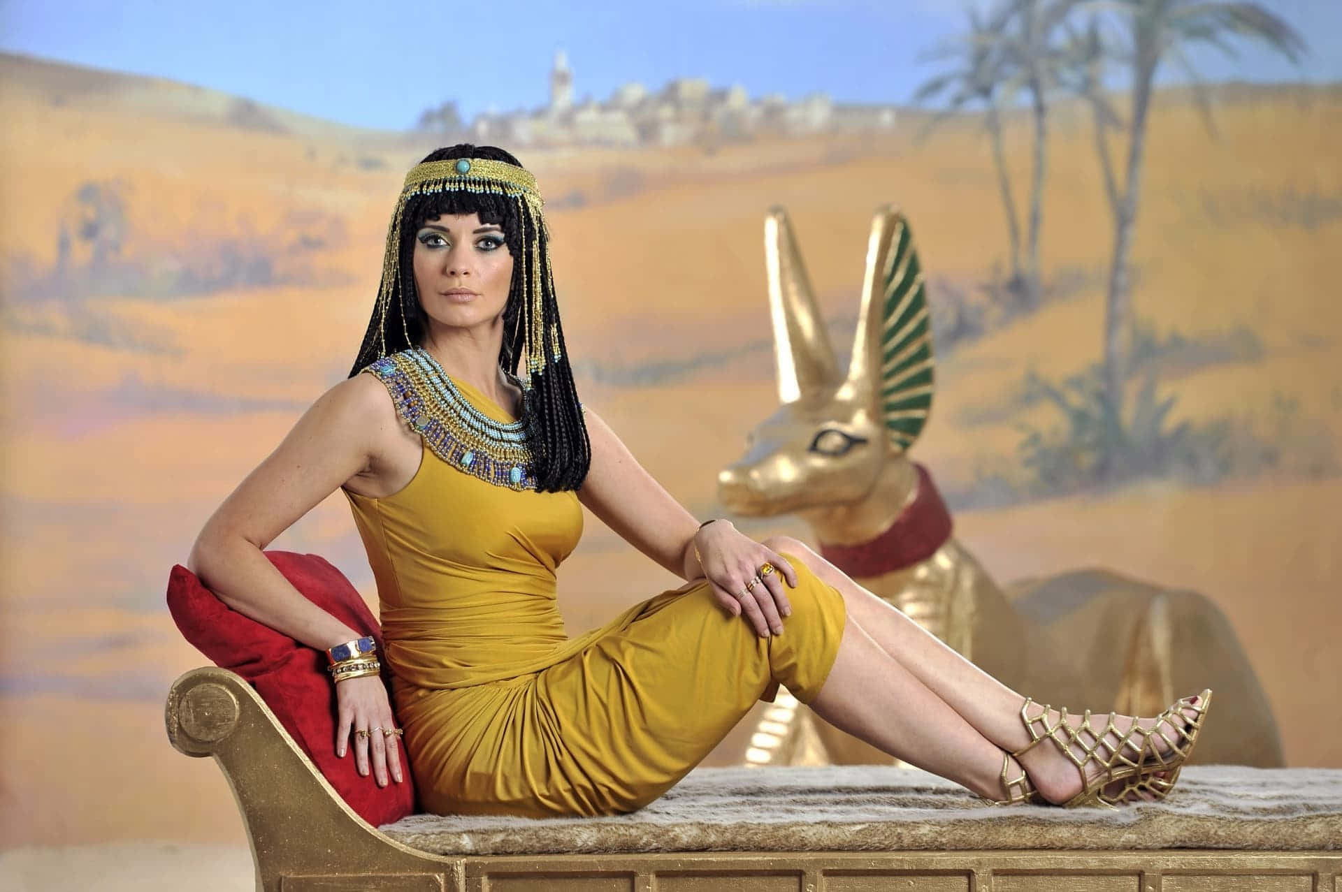 Cleopatra 桌布 壁紙 — 一位身穿金色埃及服飾的女性，在沙漠背景下威嚴地坐在金色阿努比斯雕像旁邊的寶座上。