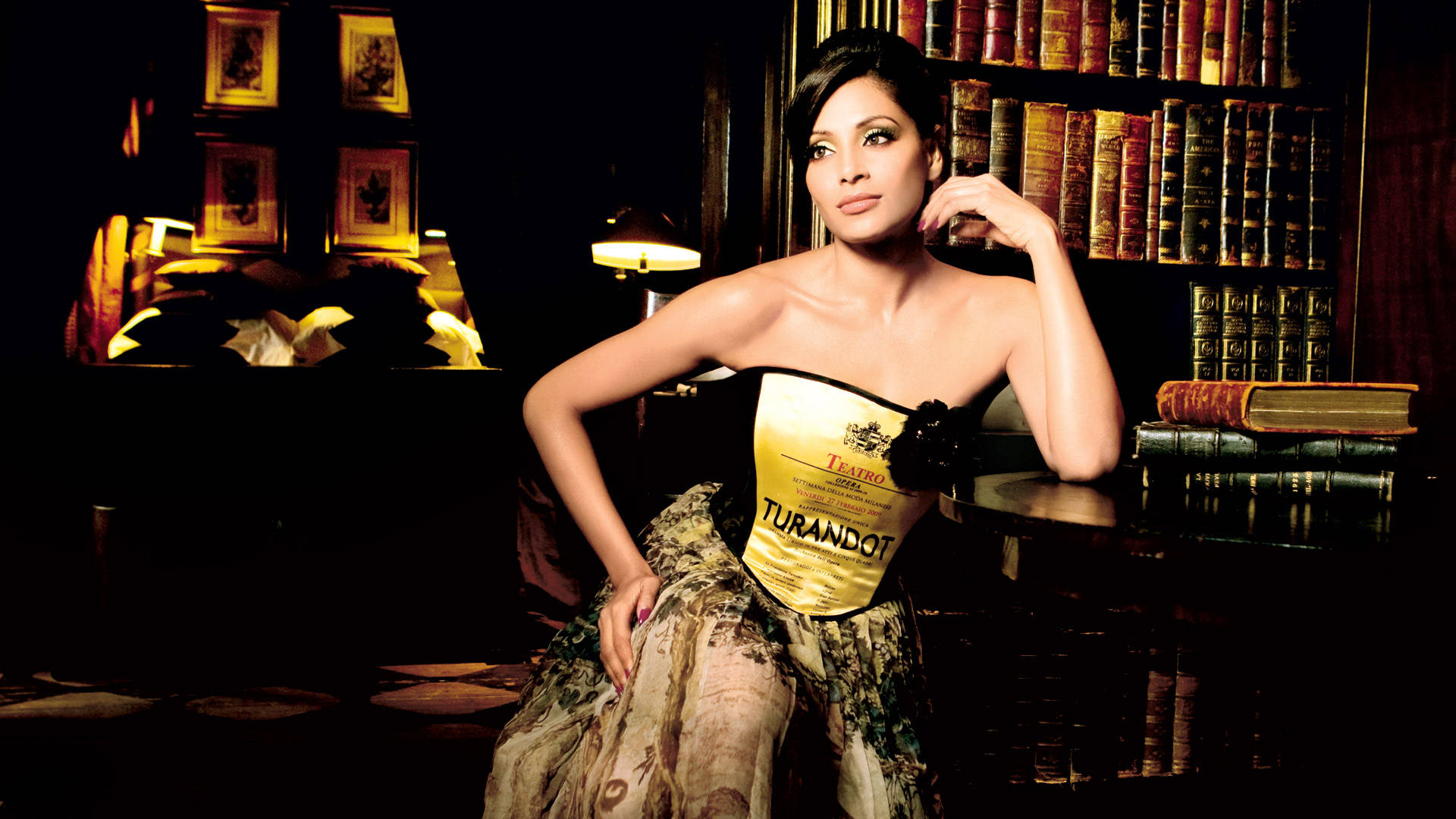Bipasha Basu 桌布 壁紙 — 一位身穿金色杜蘭朵緊身胸衣和花卉裙的女性，在光線昏暗的圖書館中，書架和檯燈旁優雅地擺姿勢。