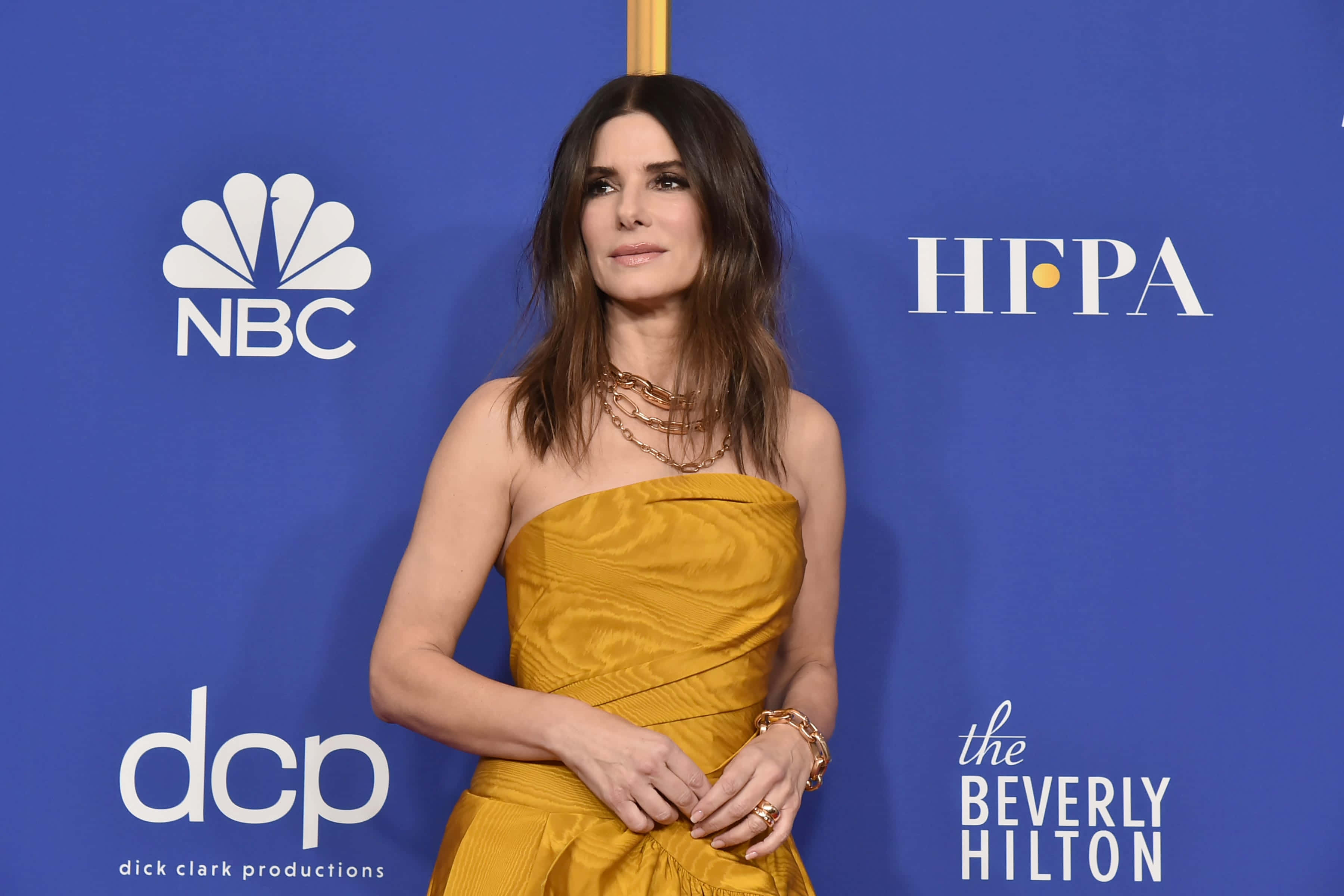 Sandra Bullock 2021 桌布 壁紙 — 一位身穿金色無肩帶連衣裙的女性在帶有標誌的藍色活動背景前自信地擺姿勢。