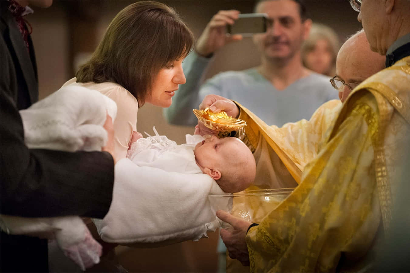 Baptism 桌布 壁紙 — 一位身穿金色祭袍的牧師在洗禮中將聖水倒在嬰兒頭上，母親則抱著裹著白布的嬰兒。