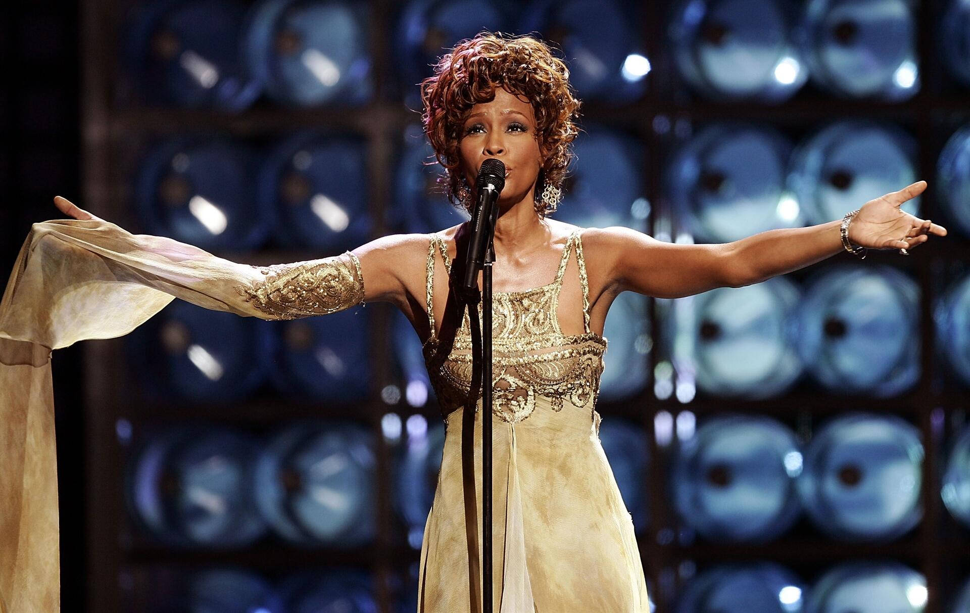 Whitney 桌布 壁紙 — 一位身穿金色禮服的魅力歌手在藍色圓形背景下張開雙臂在舞台上表演。