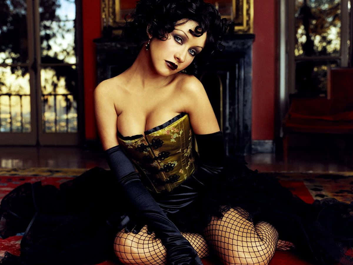 Gothic Fashion 桌布 壁紙 — 一位身穿金色緊身胸衣和黑色手套的女性，挑逗地坐在裝飾華麗的房間裡的一張圖案地毯上。