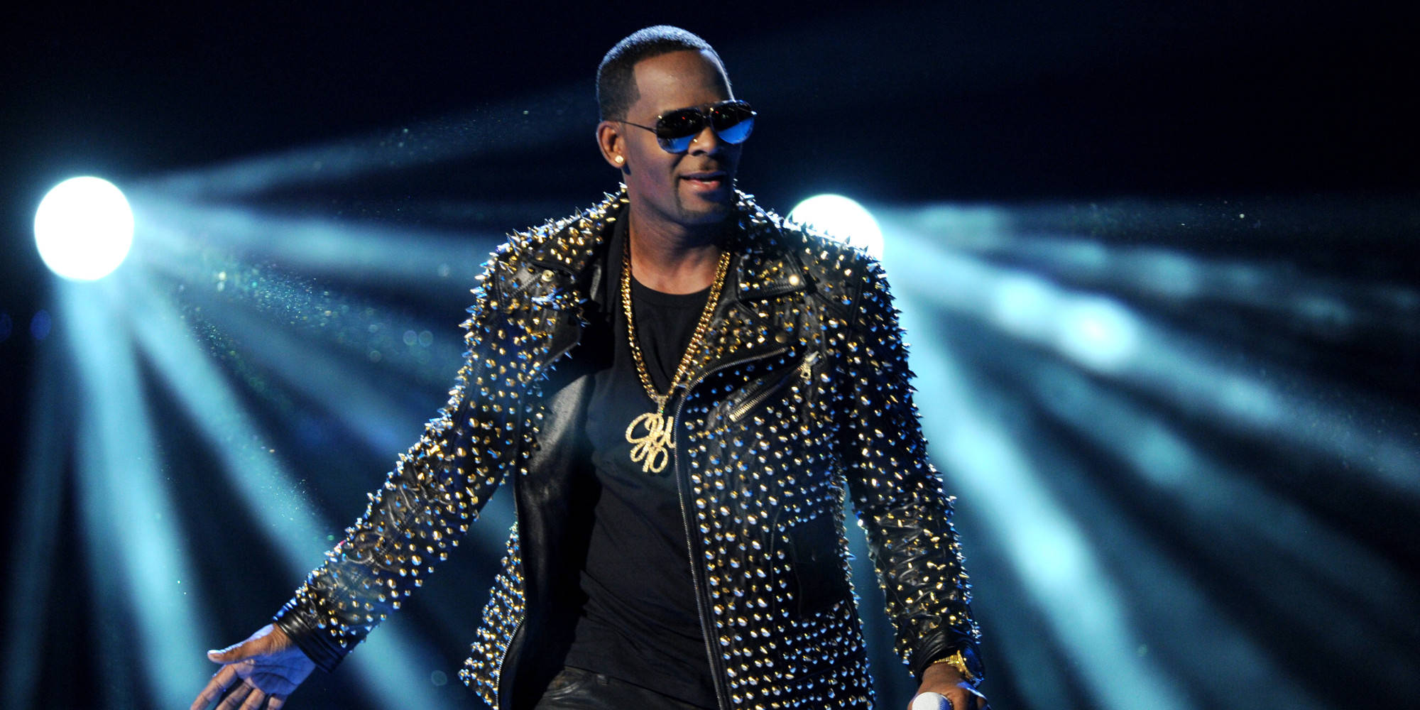 r kelly 桌布 壁紙 — 一位身穿鉚釘黑色外套和太陽眼鏡的男性表演者，在戲劇性的藍色聚光燈下掌控舞台。