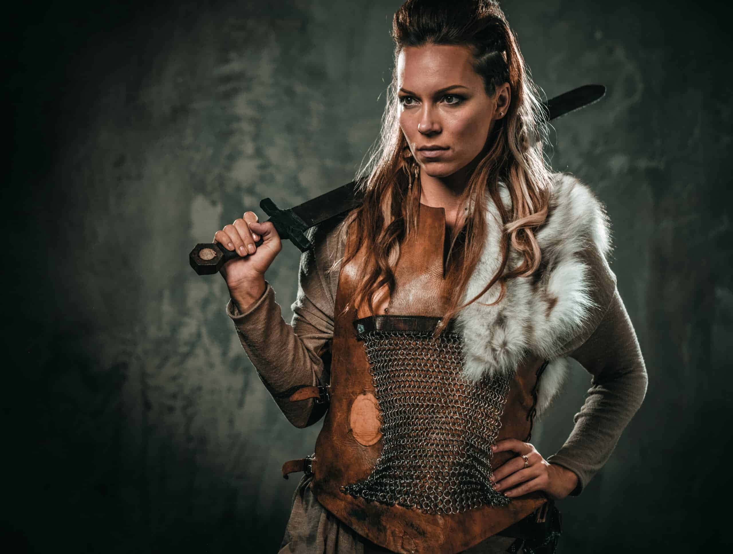 Female Viking Warriors 桌布 壁紙 — 一位身穿鎖子甲和毛皮斗篷的兇猛女戰士，在深色紋理背景上將劍扛在肩上。