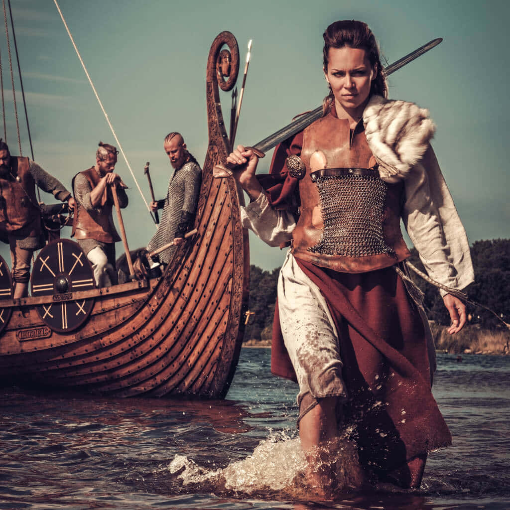Female Viking Warriors 桌布 壁紙 — 一位身穿鎖子甲和毛皮斗篷的兇猛維京女戰士，手持劍從長船踏上岸邊。