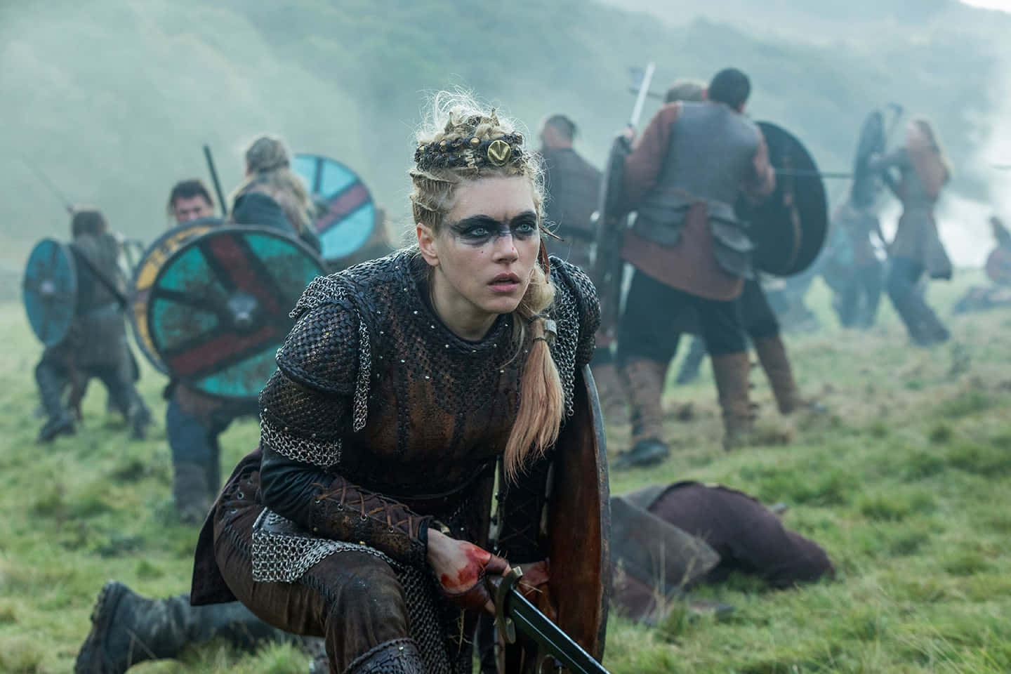 Vikings 桌布 壁紙 — 一位身穿鎖子甲的兇猛維京女戰士跪在草叢戰場上，雙手沾滿鮮血，握著劍和盾牌，身後戰士們在迷霧日光中交戰。