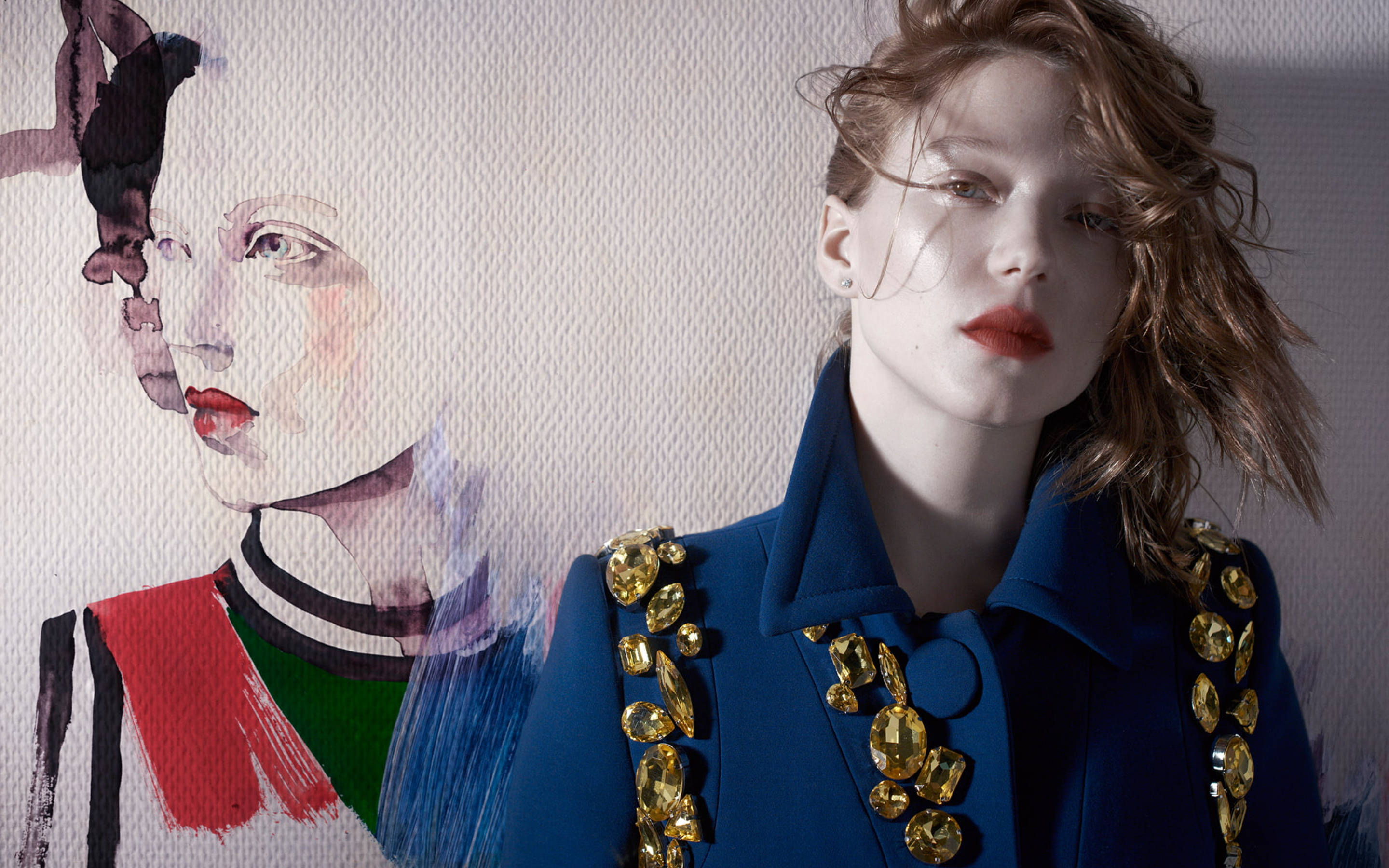 Lea Seydoux 桌布 壁紙 — 一位身穿鑲有寶石的藍色外套的女性站在有紋理的牆前，身後是另一位女性的水彩肖像。