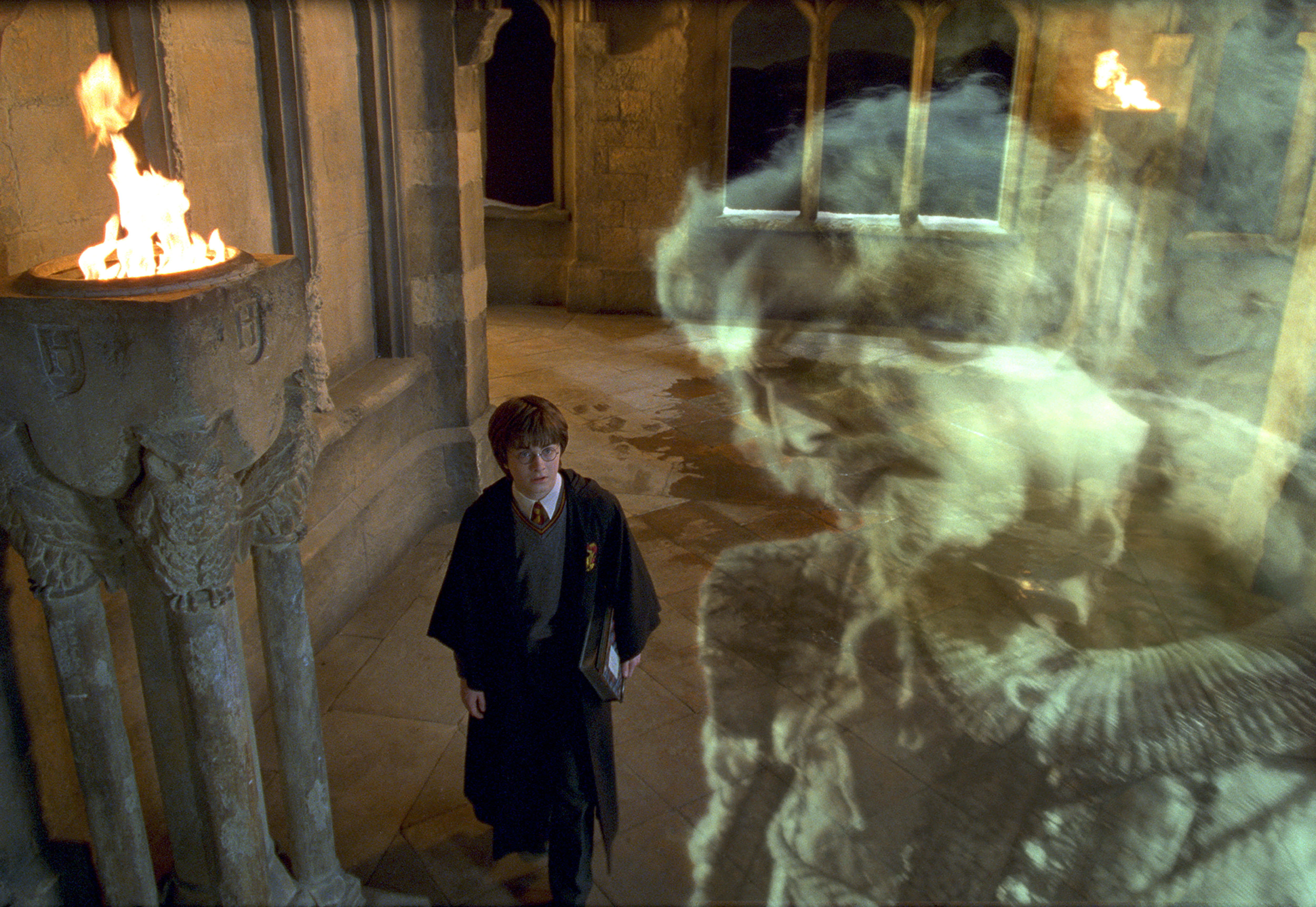 The Hogwarts Ghosts 桌布 壁紙 — 一位身穿長袍的年輕巫師走過石頭走廊，一個幽靈般的身影在他旁邊的閃爍火炬旁顯現。