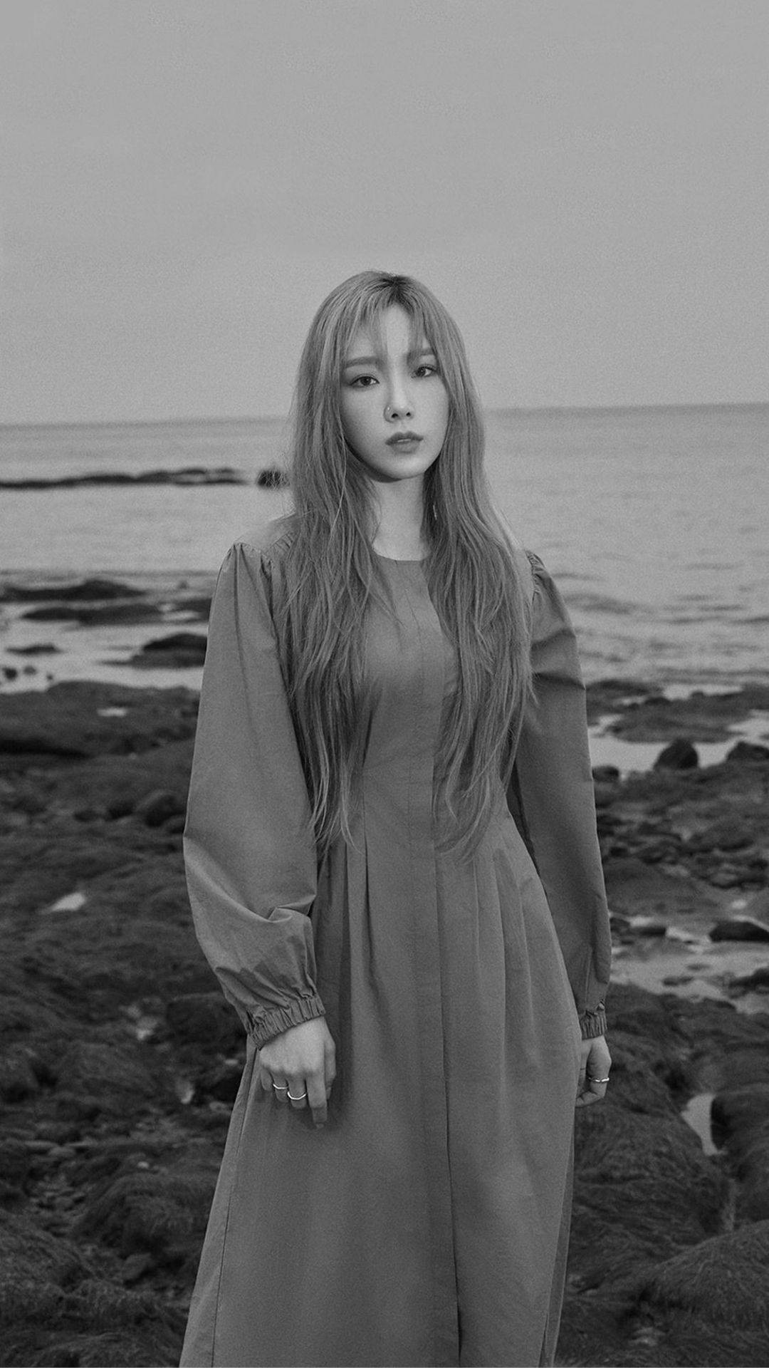 태연 桌布 壁紙 — 一位身穿長裙的女子莊嚴地站在岩石海岸線上，海浪輕輕拍打著她身後的岸邊。