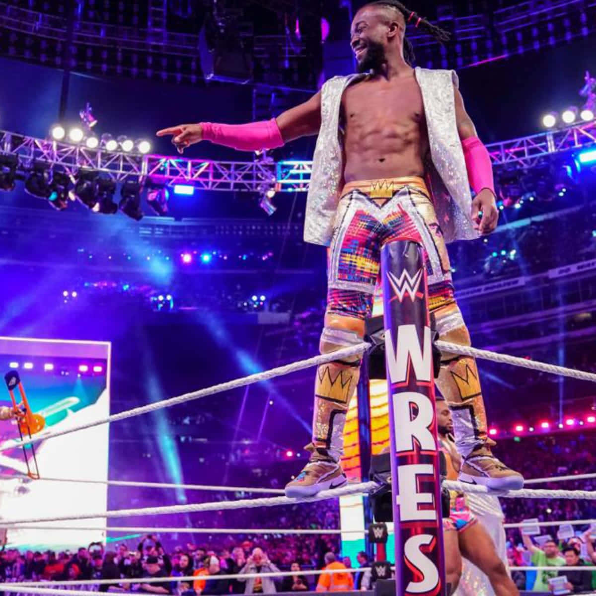 WWE 桌布 壁紙 — 一位身穿閃亮服裝的WWE摔角手，在燈光明亮的擂台角柱上勝利地站立，指向歡呼的人群。