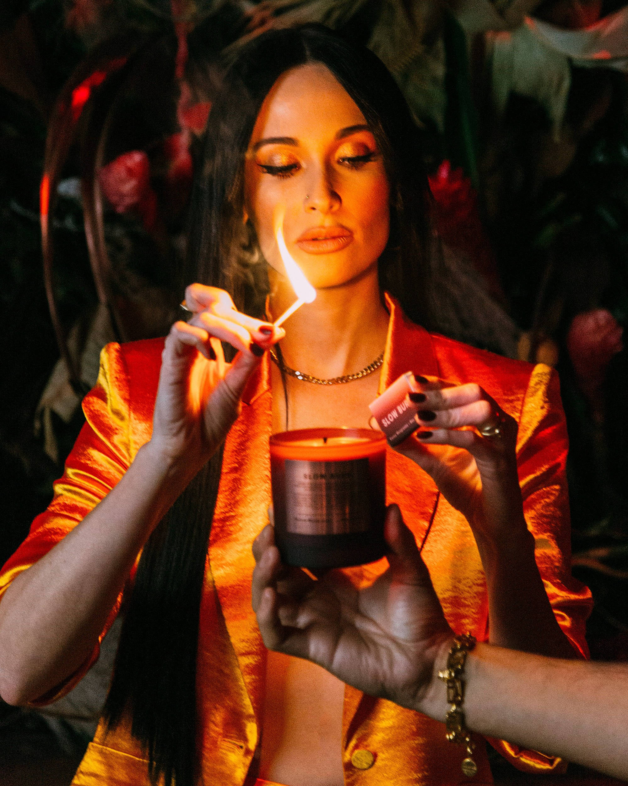 Kacey Musgraves 桌布 壁紙 — 一位身穿閃亮橙色外套的女性在光線昏暗、佈滿鮮花的房間裡用火柴點燃蠟燭。