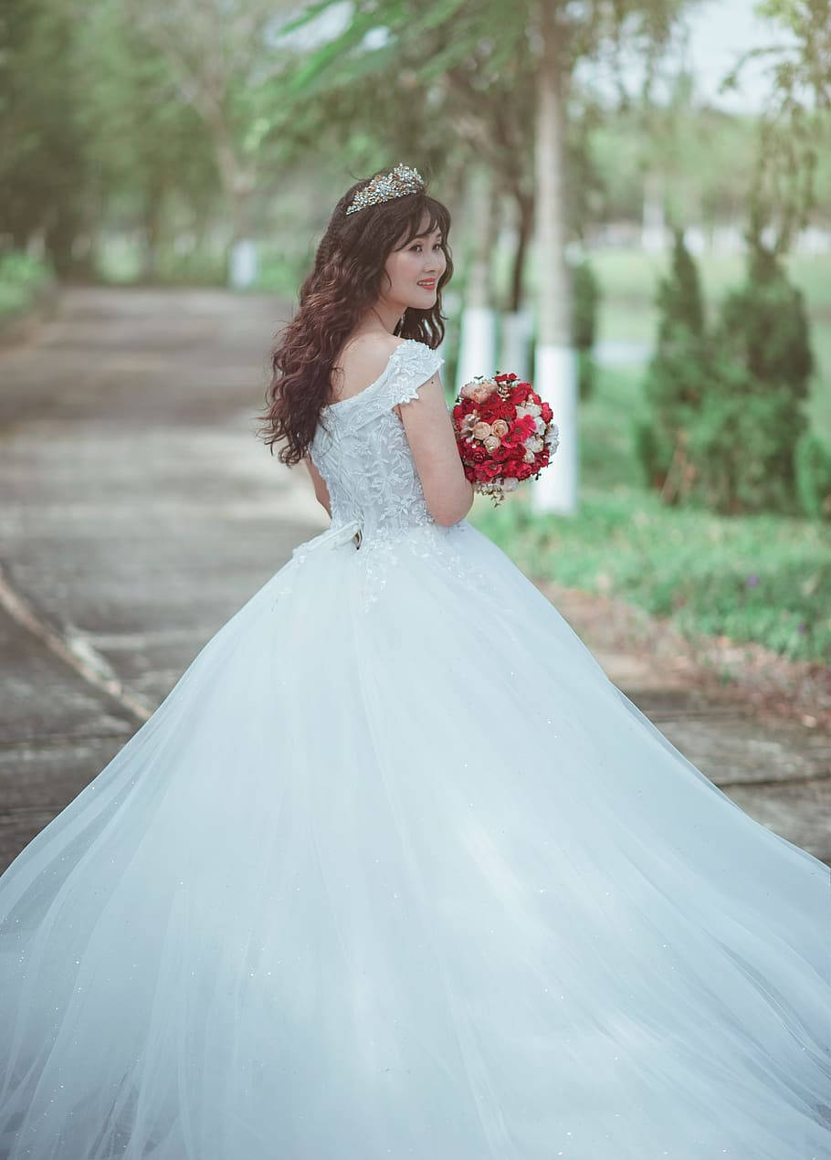 婚紗 桌布 壁紙 — 一位身穿閃亮白色婚紗、頭戴皇冠、手持紅色花束的新娘，站在林蔭小徑上，回頭微笑。